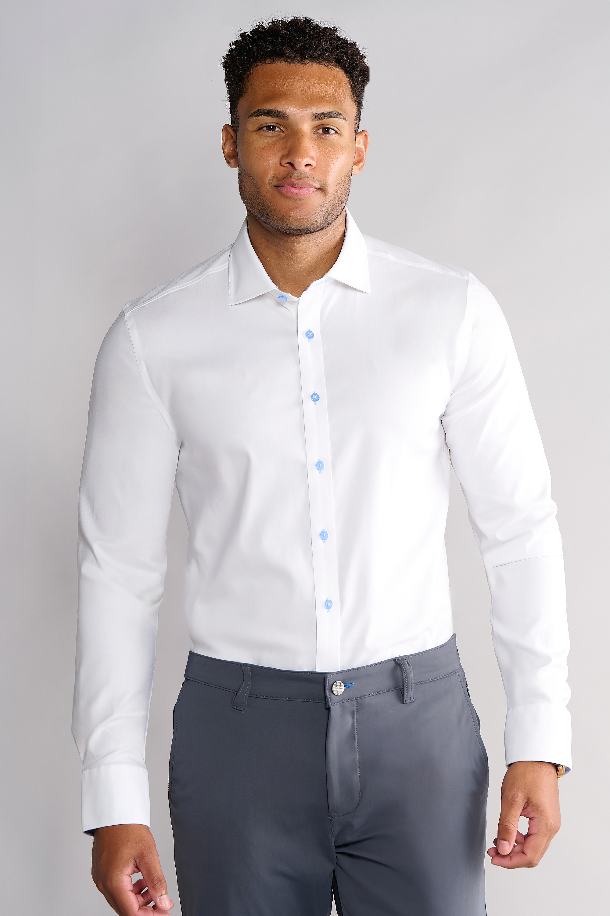 A man with short curly hair and a slight beard stands facing forward against a light gray background, wearing the Twillory Leader Contrast // Blue Prince of Wales shirt and gray dress pants, his hands relaxed by his sides.