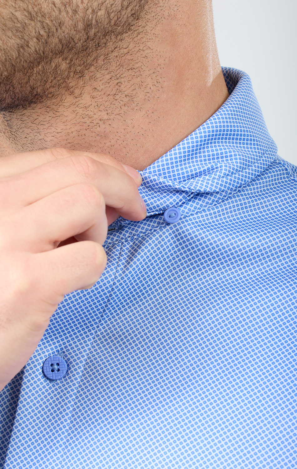 A close-up shows a person buttoning the collar of the Twillory Short Sleeve Button Down Polo Dot in Blue Diamond, focusing on the shirt’s pattern and their hand. Facial features are partially visible against a plain, blurred background.