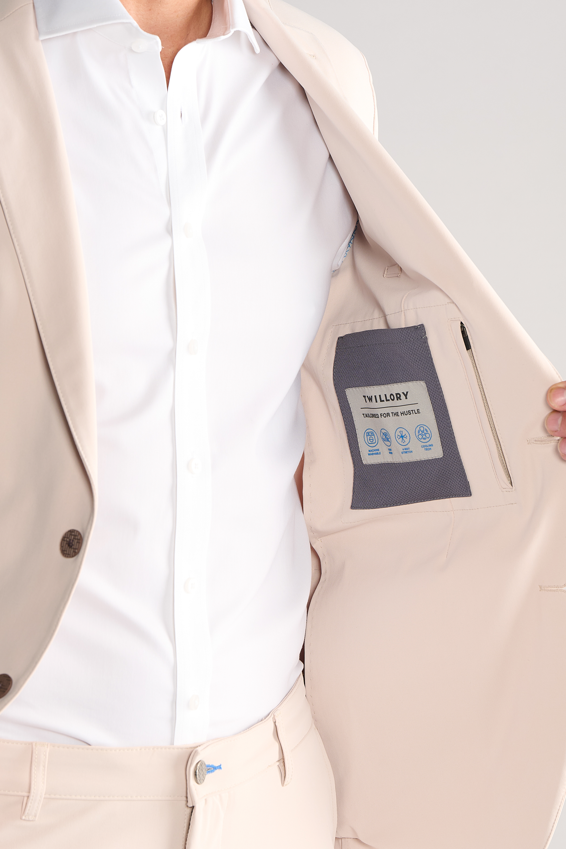 A person in a light beige Twillory Performance Blazer // Bone and white shirt holds open the jacket to reveal a labeled inner pocket with text and blue icons. Their head is out of frame against a plain light gray background.