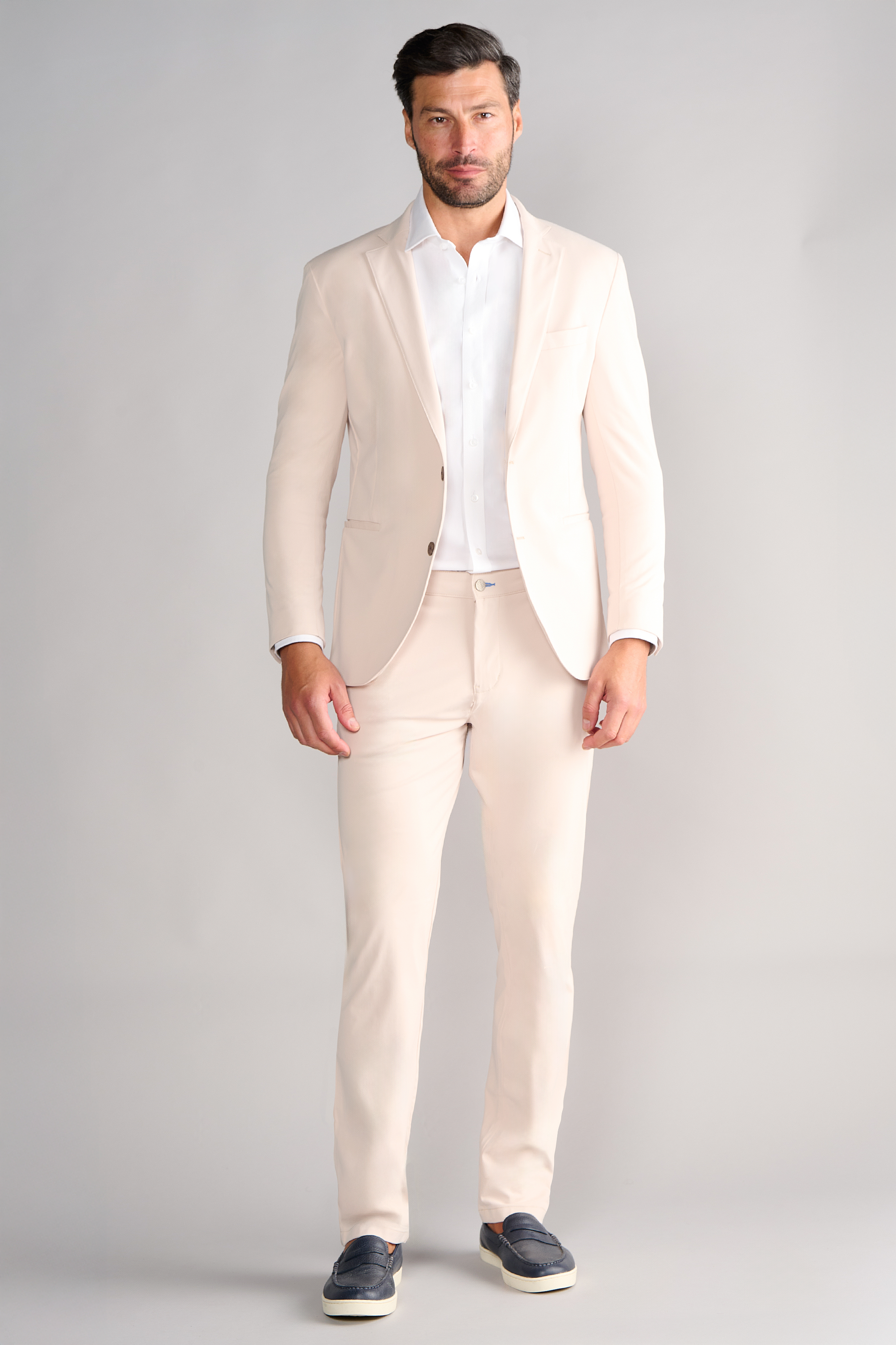 A man with short dark hair and a trimmed beard stands facing forward against a light grey background, wearing Twillory’s Performance Blazer // Bone over a white dress shirt and black slip-on shoes, hands by his sides.