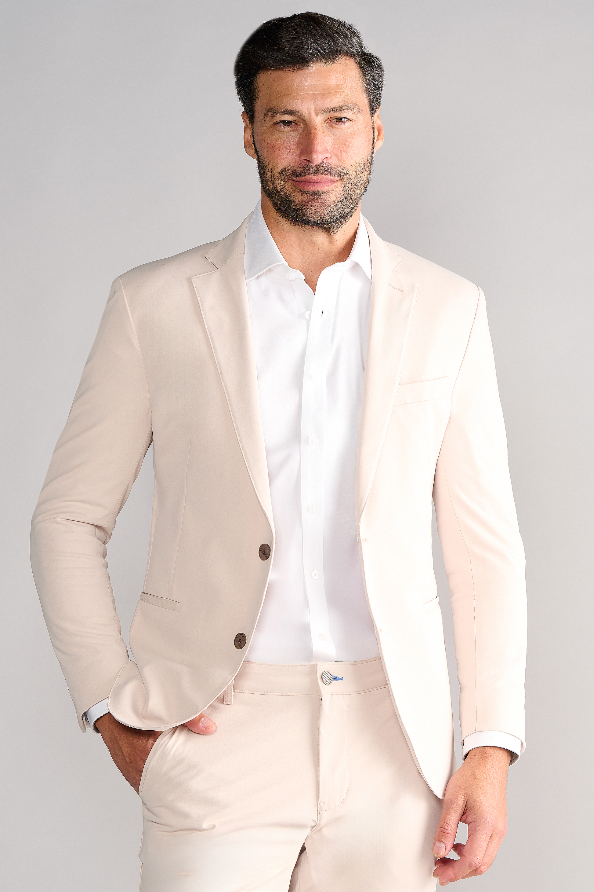 A man with short dark hair and a beard models Twillory's Performance Blazer // Bone over a white dress shirt, standing with one hand in his pocket and smiling slightly against a light gray backdrop.