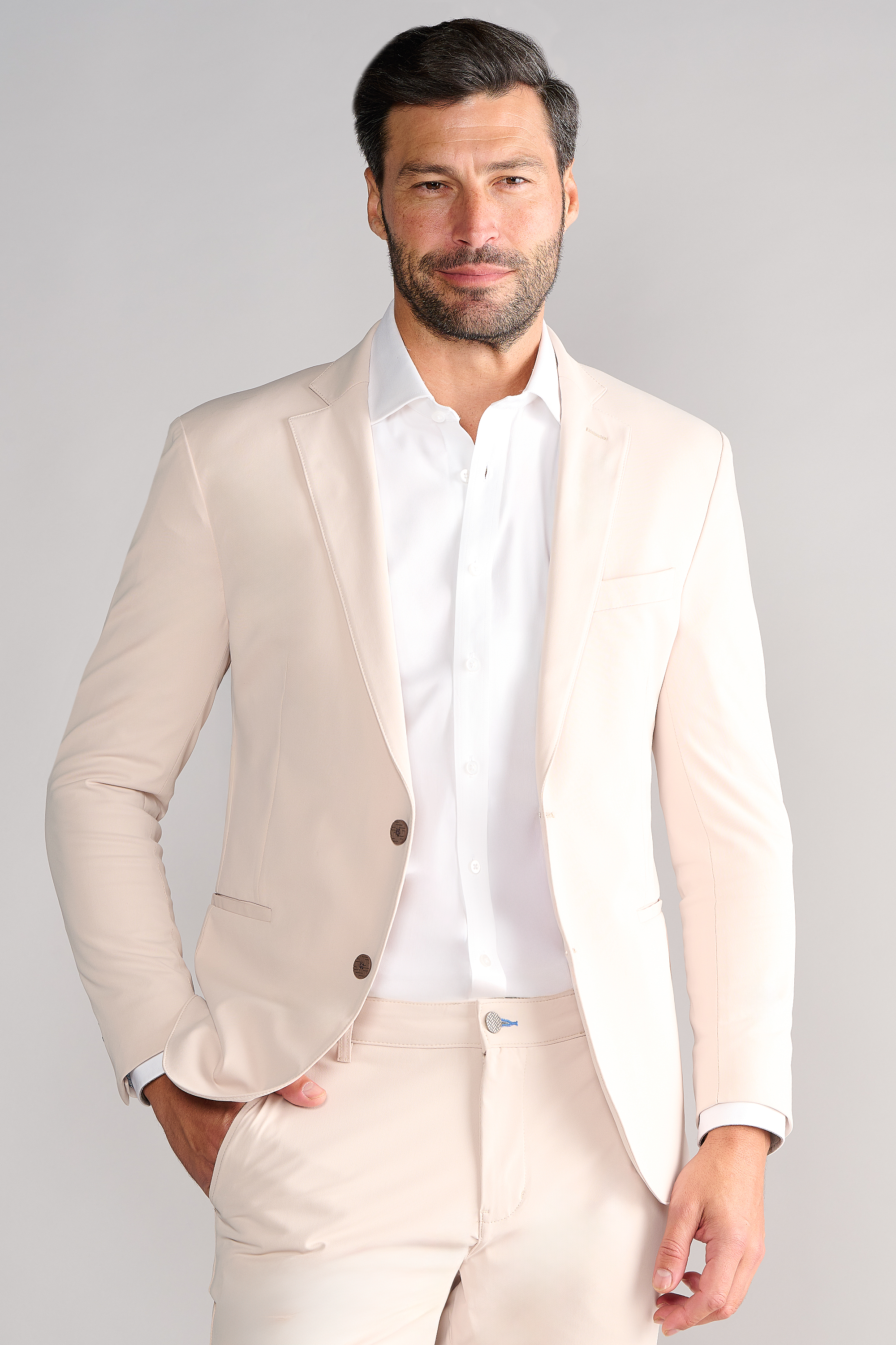 A man with short dark hair and a beard models Twillory's Performance Blazer // Bone over a white dress shirt, standing with one hand in his pocket and smiling slightly against a light gray backdrop.
