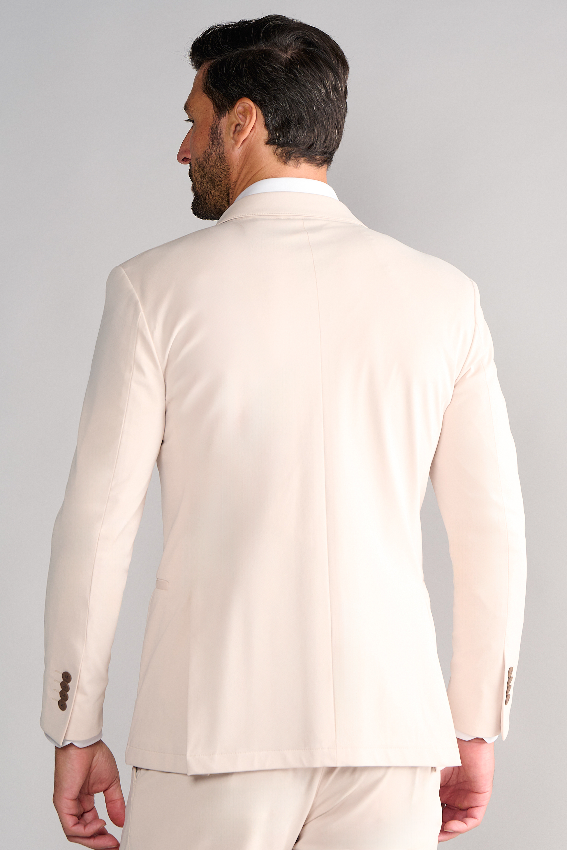 A man faces away from the camera in a Twillory Performance Blazer // Bone with matching trousers. The light beige blazer features brown cuff buttons and a center back seam, set against a neutral light gray background.