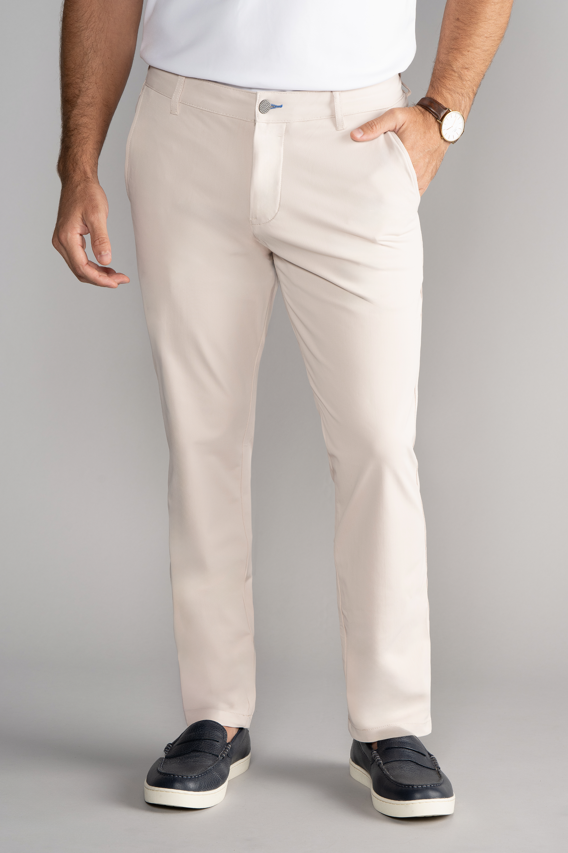 A man stands against a gray background wearing Twillory Performance Pants in Bone, a tucked-in white shirt, brown wristwatch, and dark blue loafers with white soles. His left hand is in his pocket while his right arm hangs by his side.
