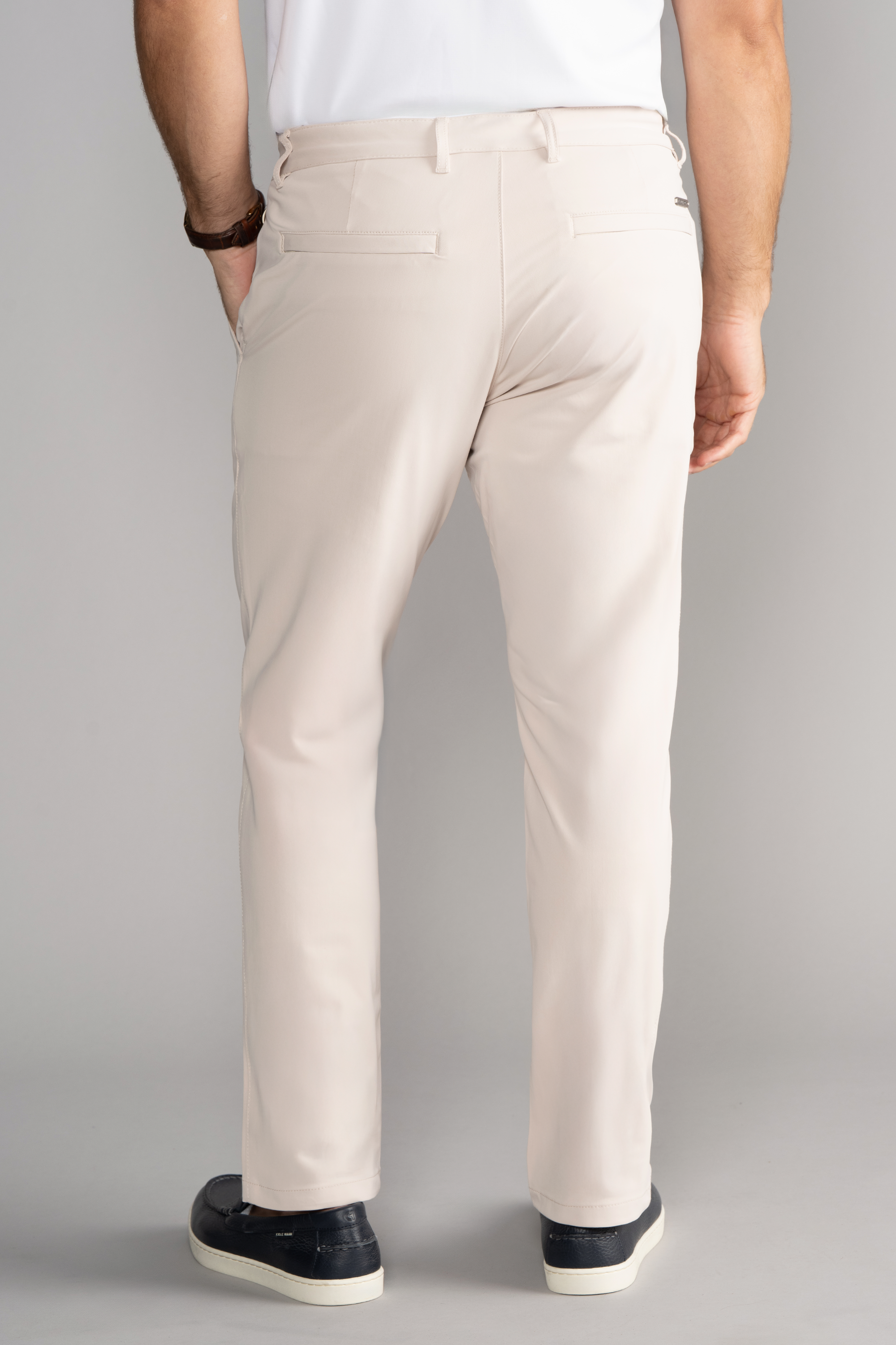 A man faces away, wearing Twillory Performance Pants // BONE, a white short-sleeve shirt, and black low-top sneakers with white soles. His left hand is by his side, a dark wristband on his right wrist. The background is plain gray.