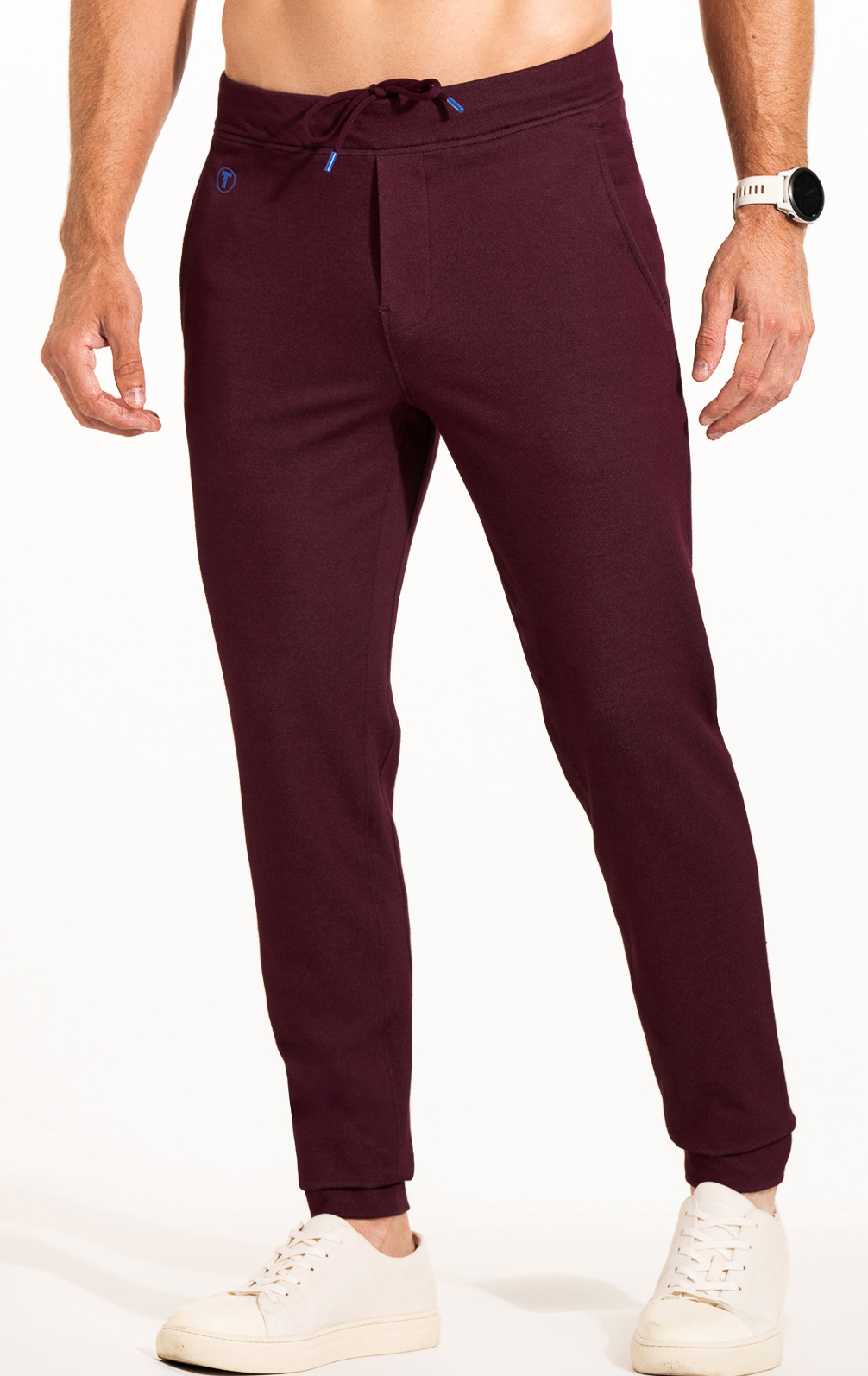 A man models Twillory's Athlounger Jogger—maroon comfort-fabric pants with a drawstring waist and side-zip pocket—paired with white sneakers and a wristwatch, standing against a white background. His upper body is not visible; hands relaxed by his sides.