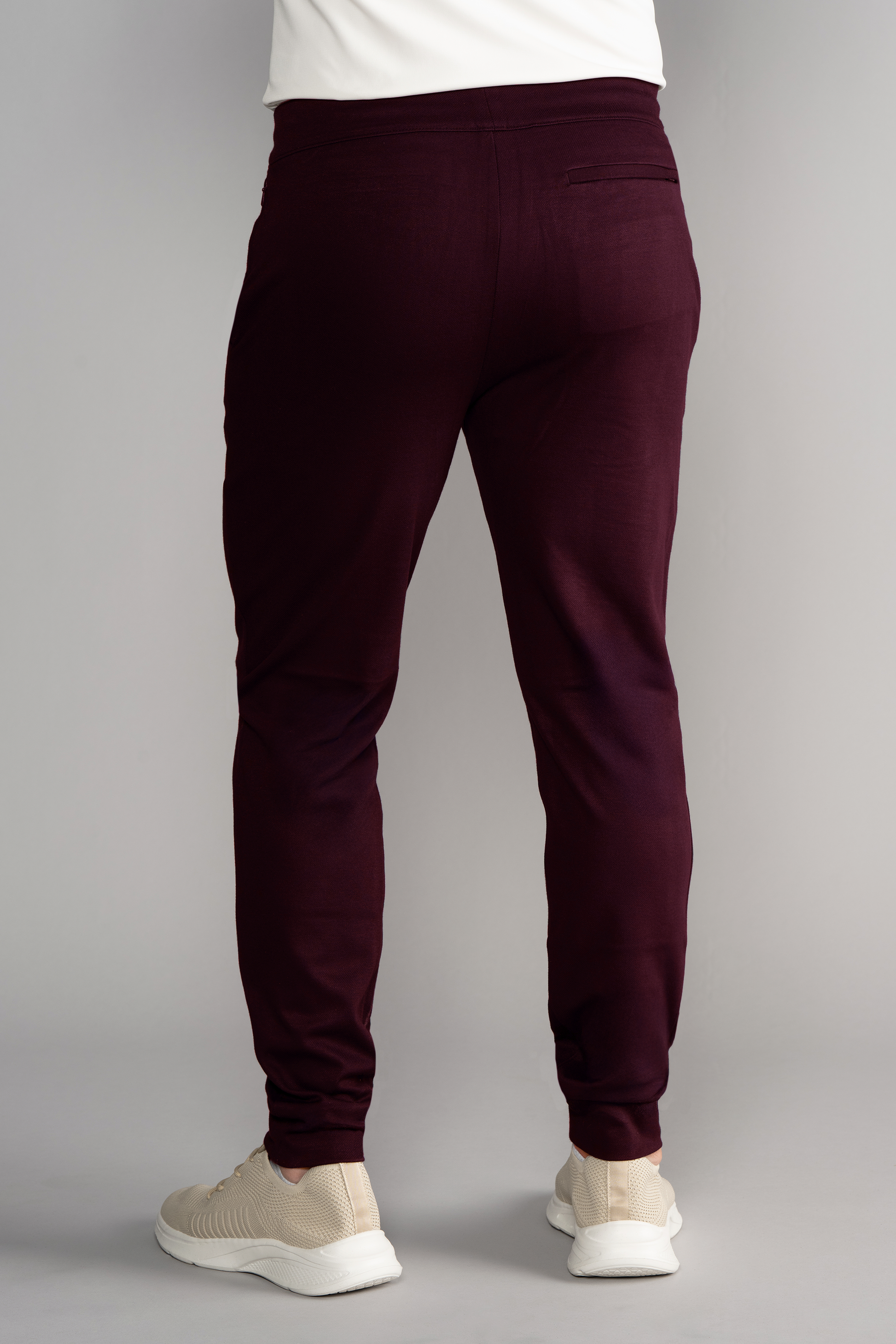 A person models Twillory’s Athlounger Jogger, slim-fit burgundy pants made from comfort fabric with a single back pocket and paired with white sneakers. The plain light gray background shows only the lower half of their body.