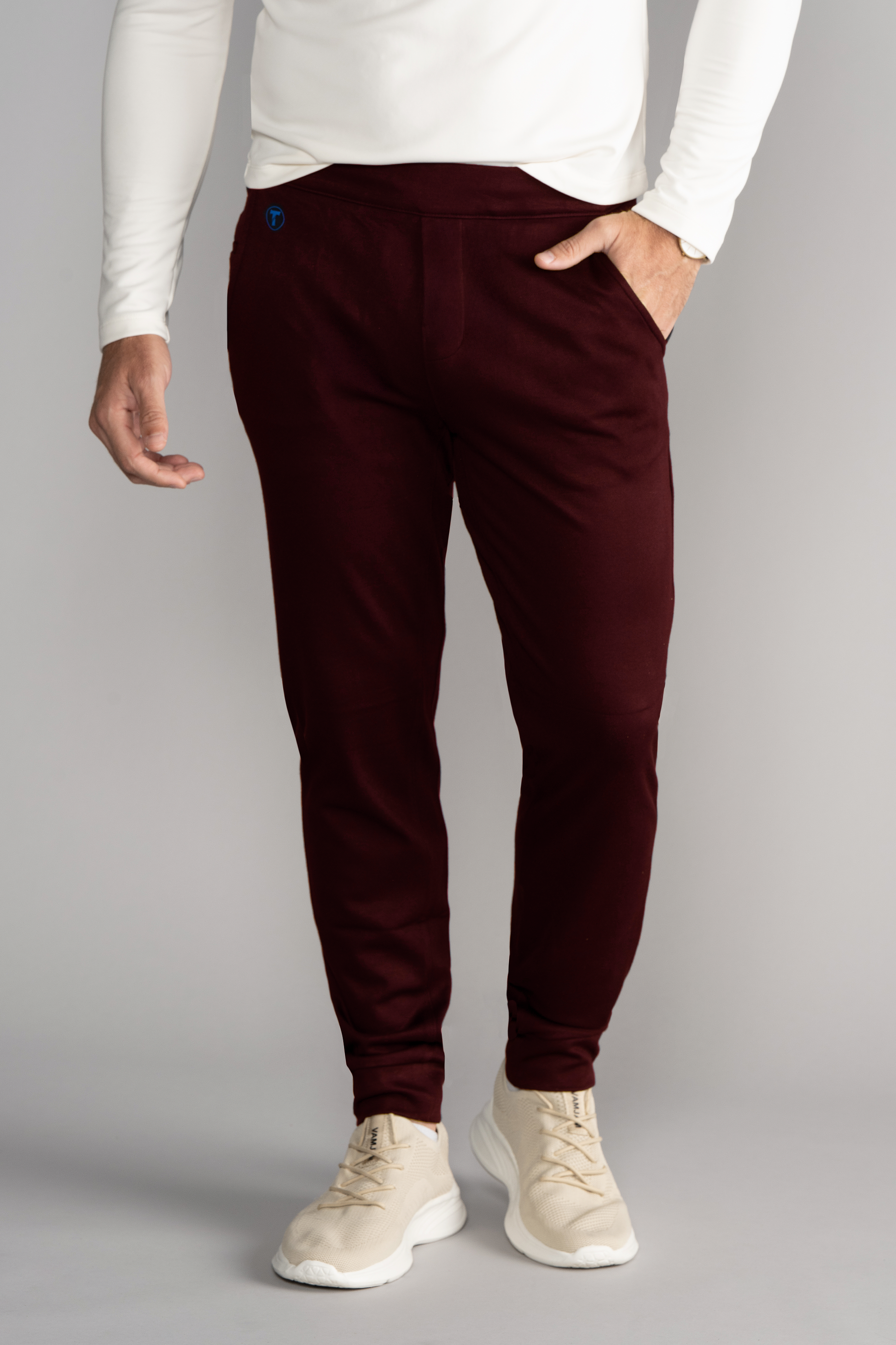 A person wears a long-sleeve white shirt, Twillory Athlounger Jogger in burgundy with a blue logo on the left hip and side-zip pocket, plus beige sneakers. They're standing against a plain light gray background, left hand in their pocket.