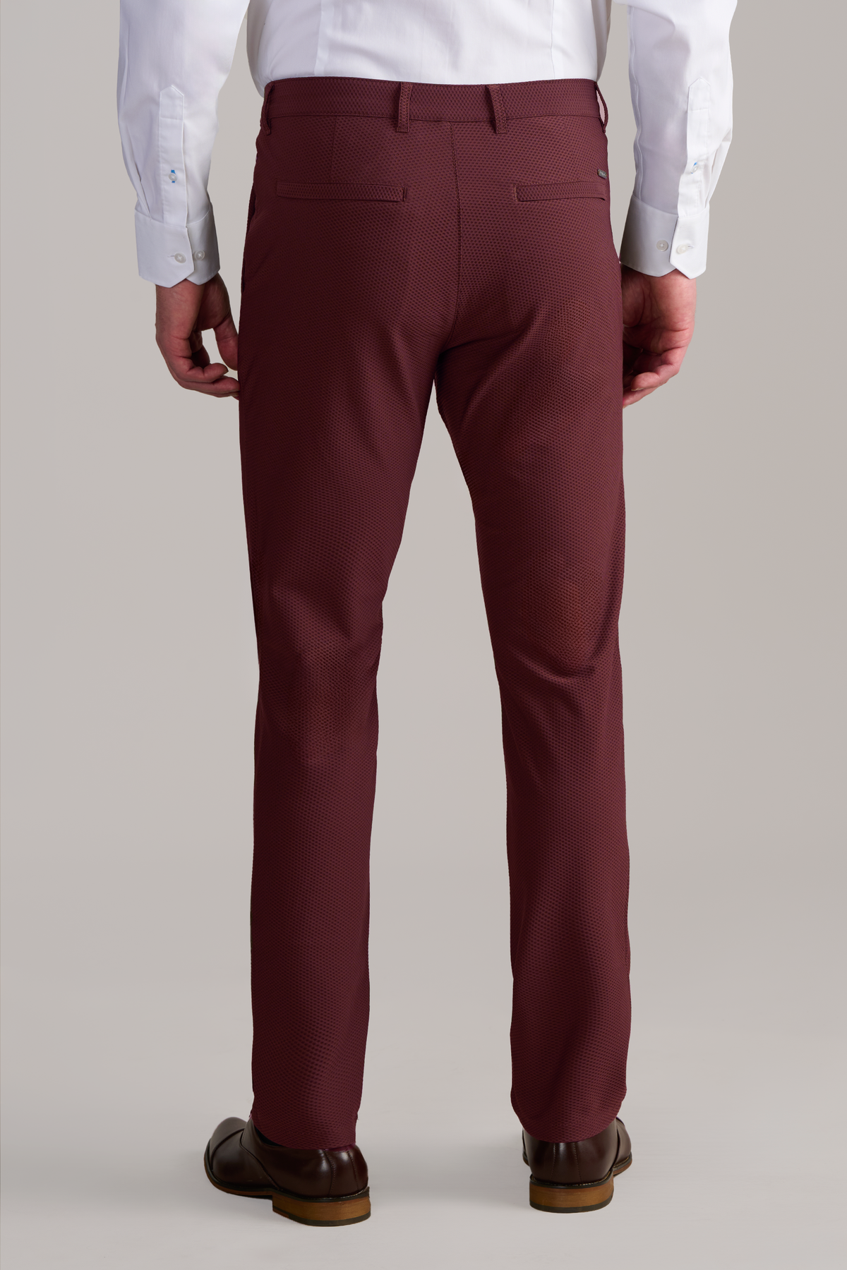 A person stands facing away, wearing Twillory’s AIR Pants in Burgundy with a textured pattern, two back welt pockets, and a white button-down shirt. The stretch pants offer all-day comfort. Brown dress shoes complete the look against a plain light background.
