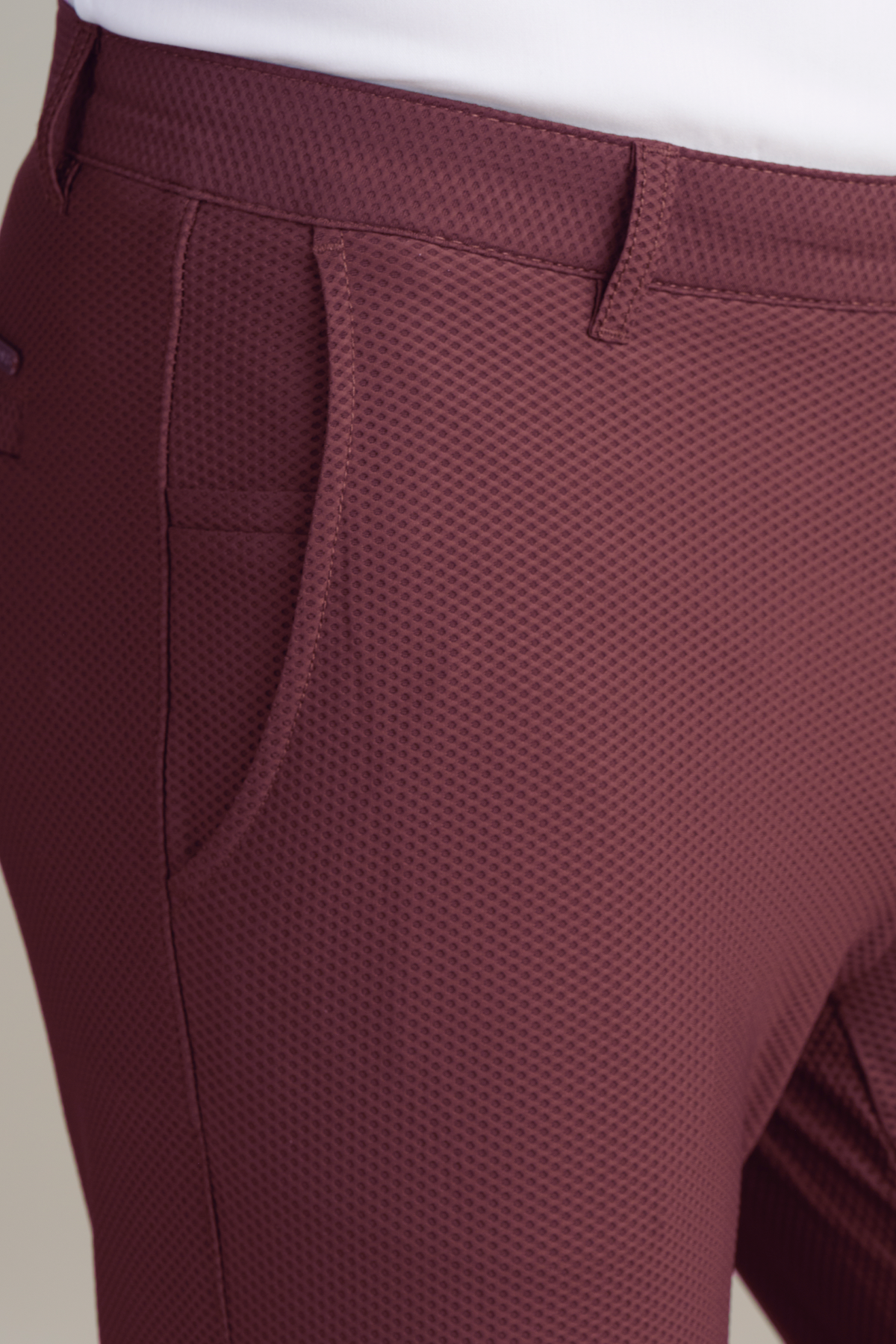 Close-up of a person wearing Twillory's AIR Pants // Burgundy, featuring a textured geometric pattern, visible side pocket, and belt loop. The look is complemented by the hem of a white top under soft lighting.