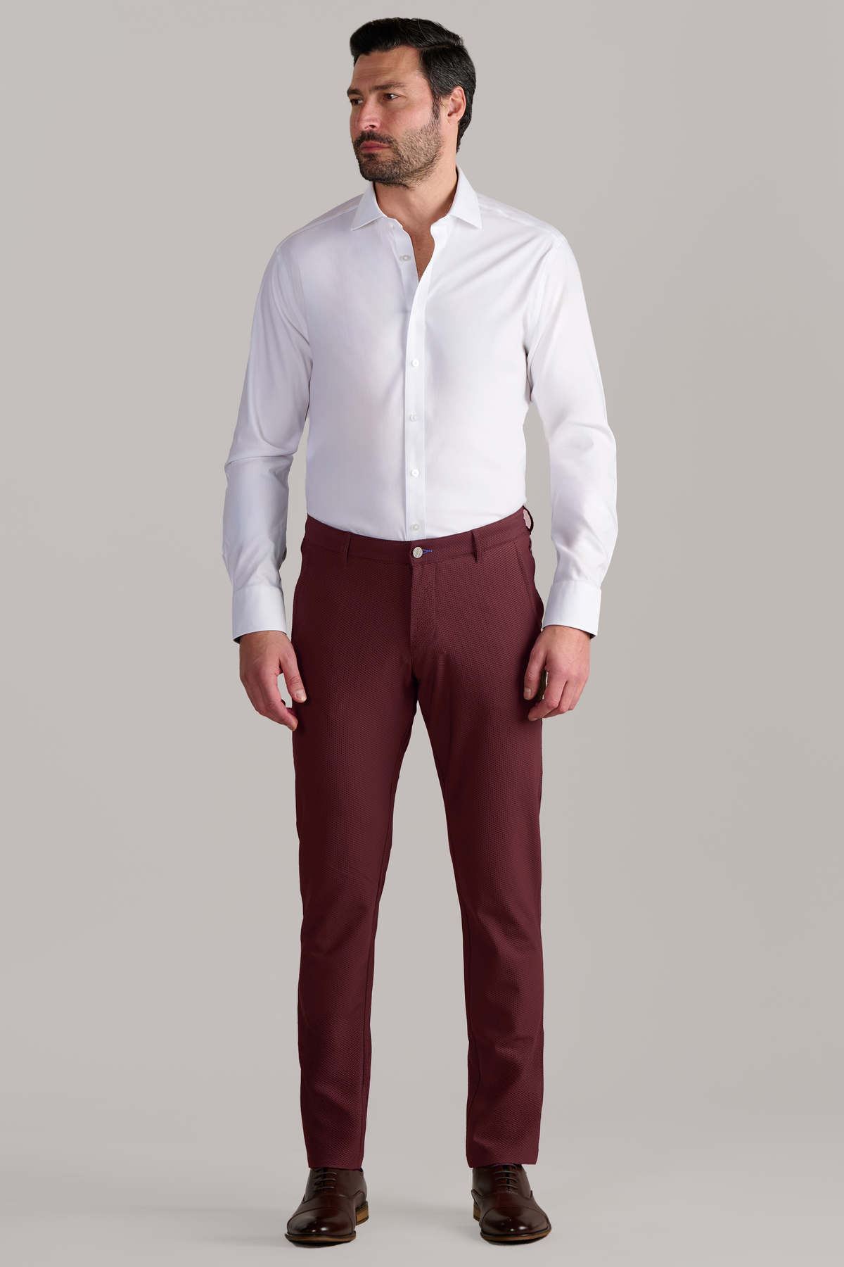 A man with short dark hair and a beard stands against a light gray background, wearing Twillory AIR Pants in Burgundy with a white dress shirt and brown shoes. His arms rest at his sides in a relaxed pose.