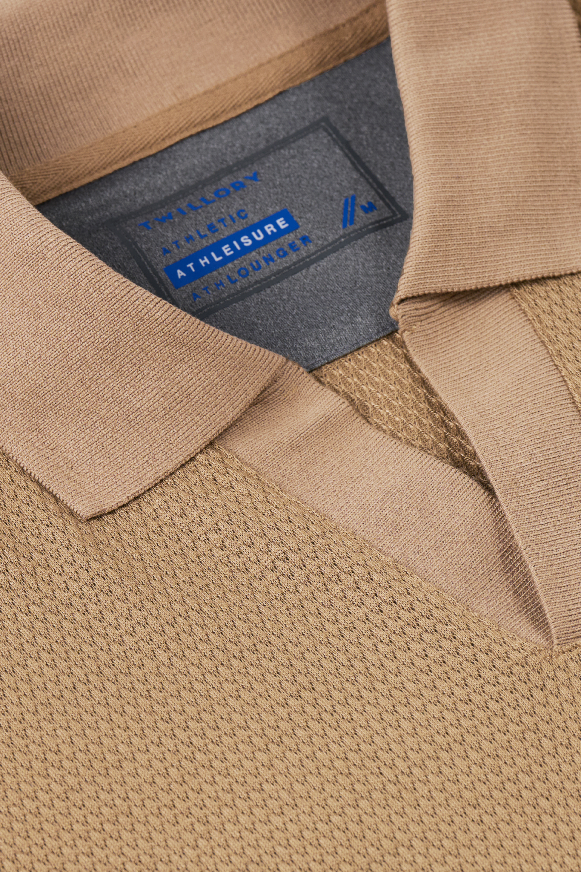 A close-up of Twillory’s Long Sleeve Sweater Polo Solid in Camel, featuring a soft collar and performance knit fabric. The navy and white inside label reads “ATHLEISURE,” emphasizing its wrinkle-free design.