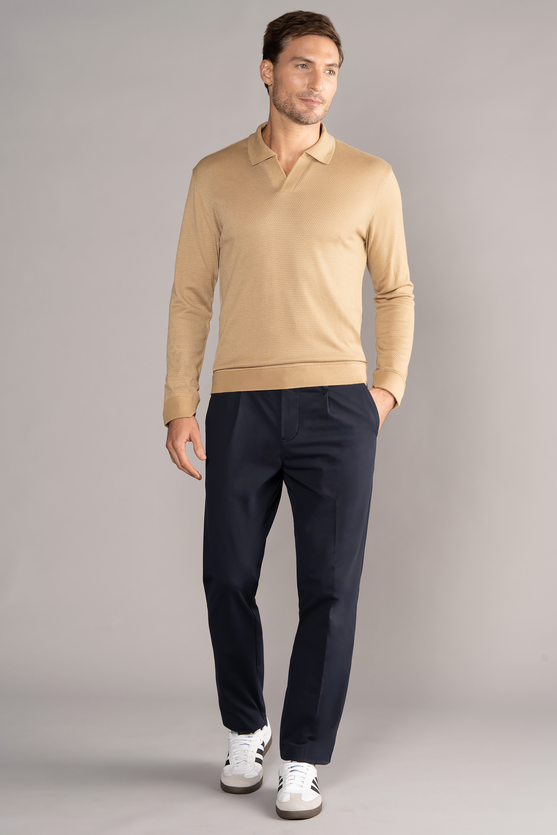 A man with short brown hair wears the Twillory Long Sleeve Sweater Polo Solid in Camel, paired with dark navy trousers and white sneakers. He stands casually against a light gray background, looking relaxed and slightly to the side.