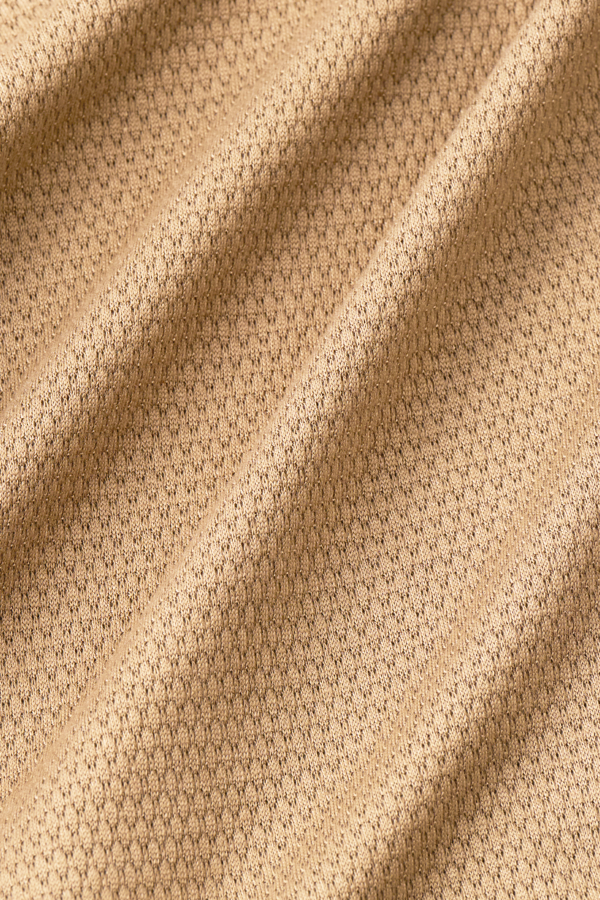Close-up of Twillory's Long Sleeve Sweater Polo Solid in Camel: light brown performance knit with a small hexagonal pattern. Soft folds and gentle shadows highlight its smooth, wrinkle-free texture and uniform woven surface.