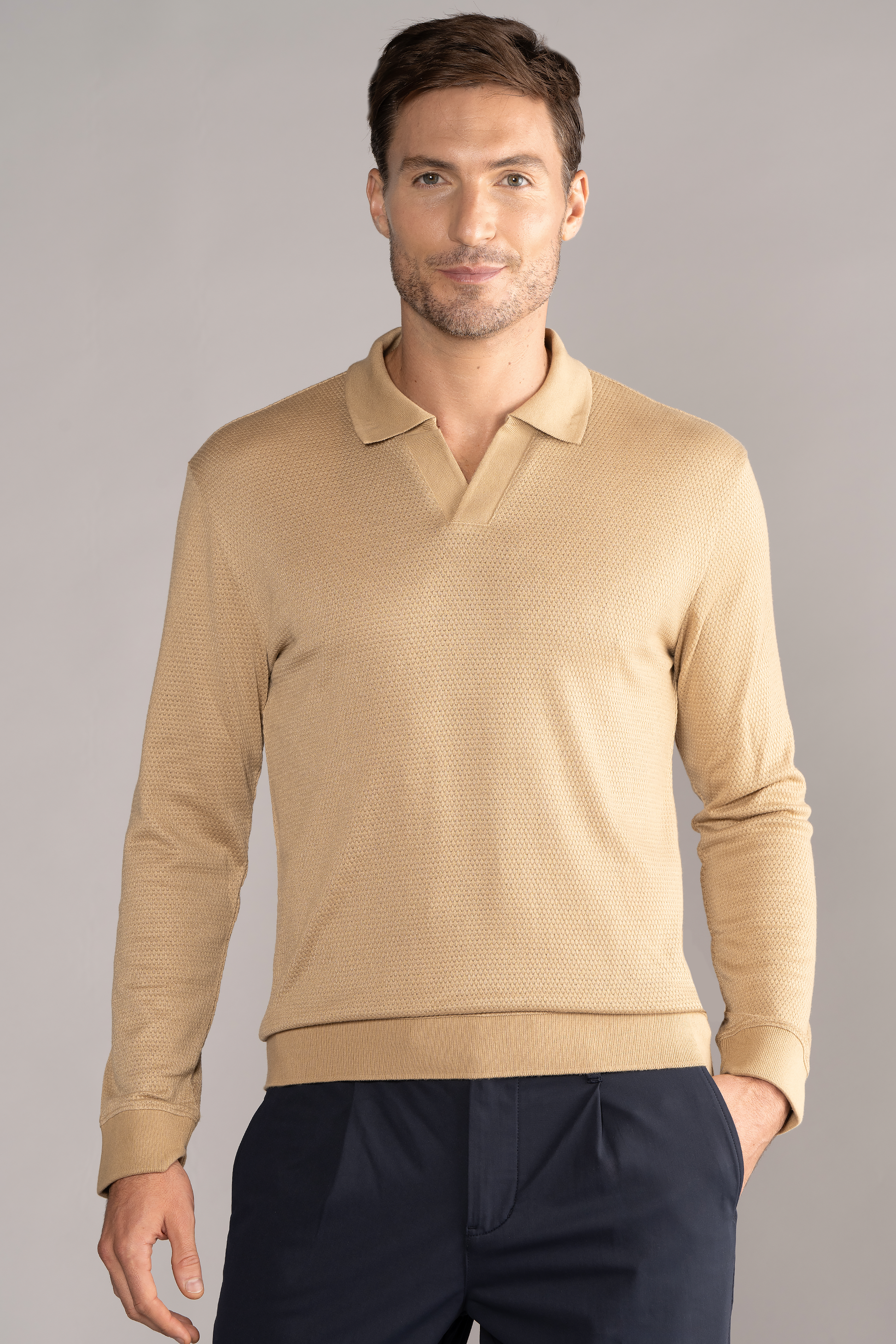 A man with short brown hair and light skin stands facing forward against a gray background, wearing the Twillory Long Sleeve Sweater Polo Solid in Camel—featuring wrinkle-free knit fabric, collar, no visible buttons—paired with dark navy pants.