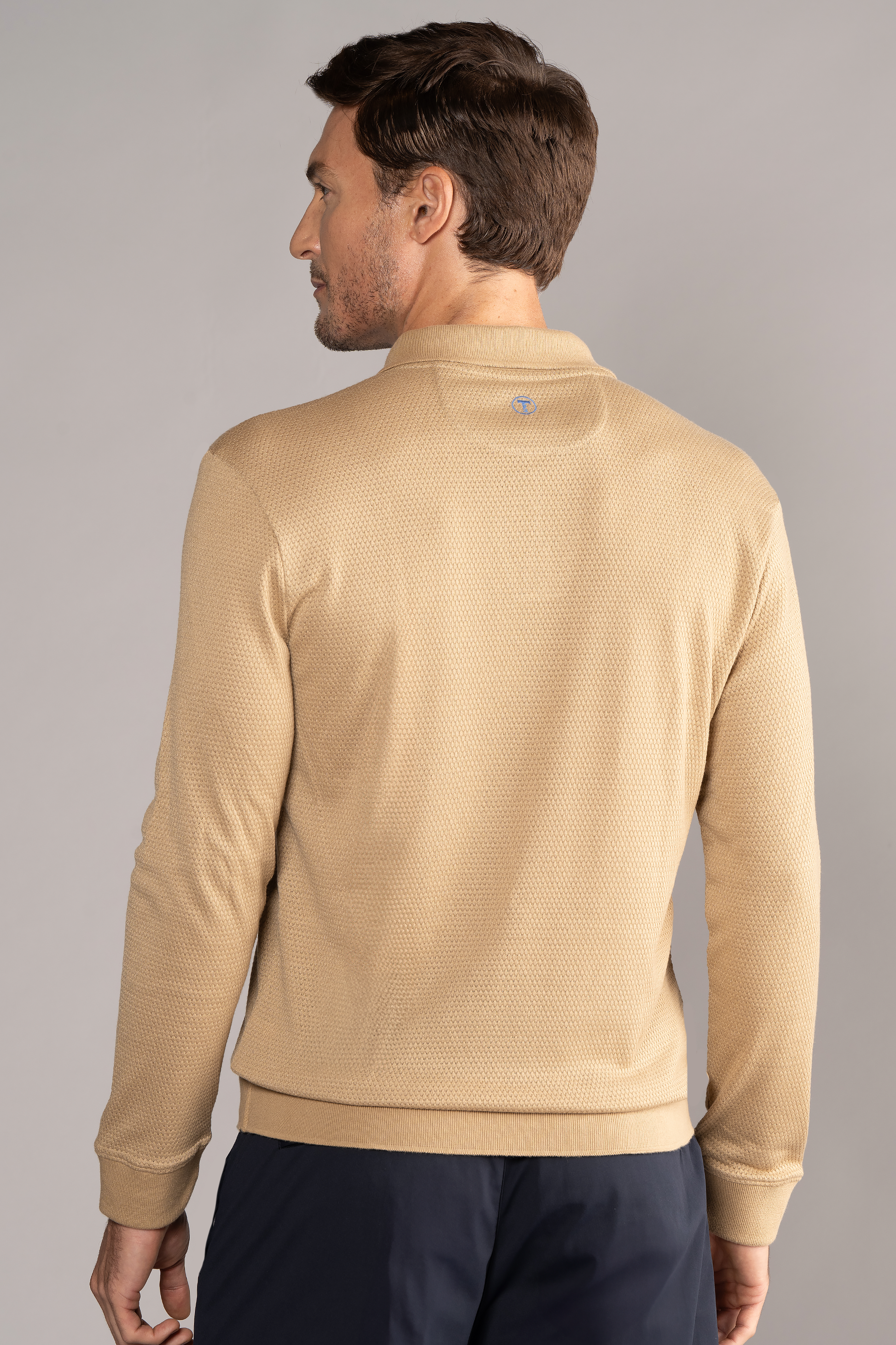 A man with short brown hair is seen from behind wearing Twillory's Long Sleeve Sweater Polo Solid in Camel and dark navy pants against a plain light gray background, his head slightly turned to the left.