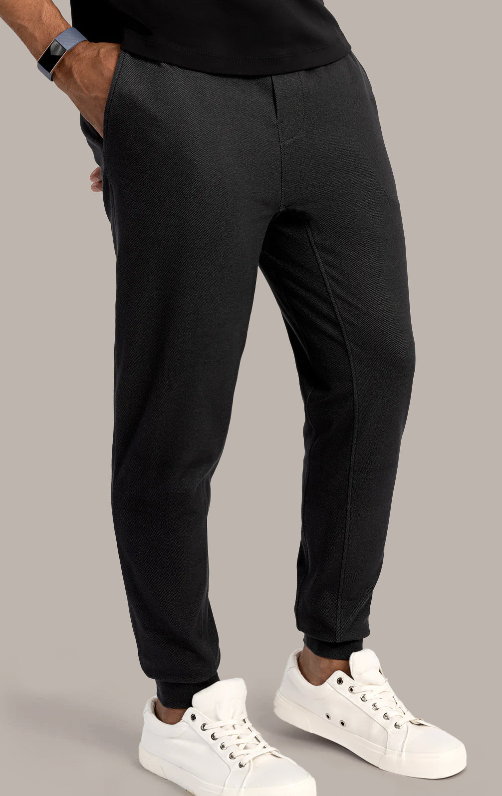 A person wears Twillory Athlounger Jogger in black with a side-zip pocket, paired with a black shirt, white sneakers, and a black smartwatch. Standing hands in pockets against a beige background; only the lower half is shown.