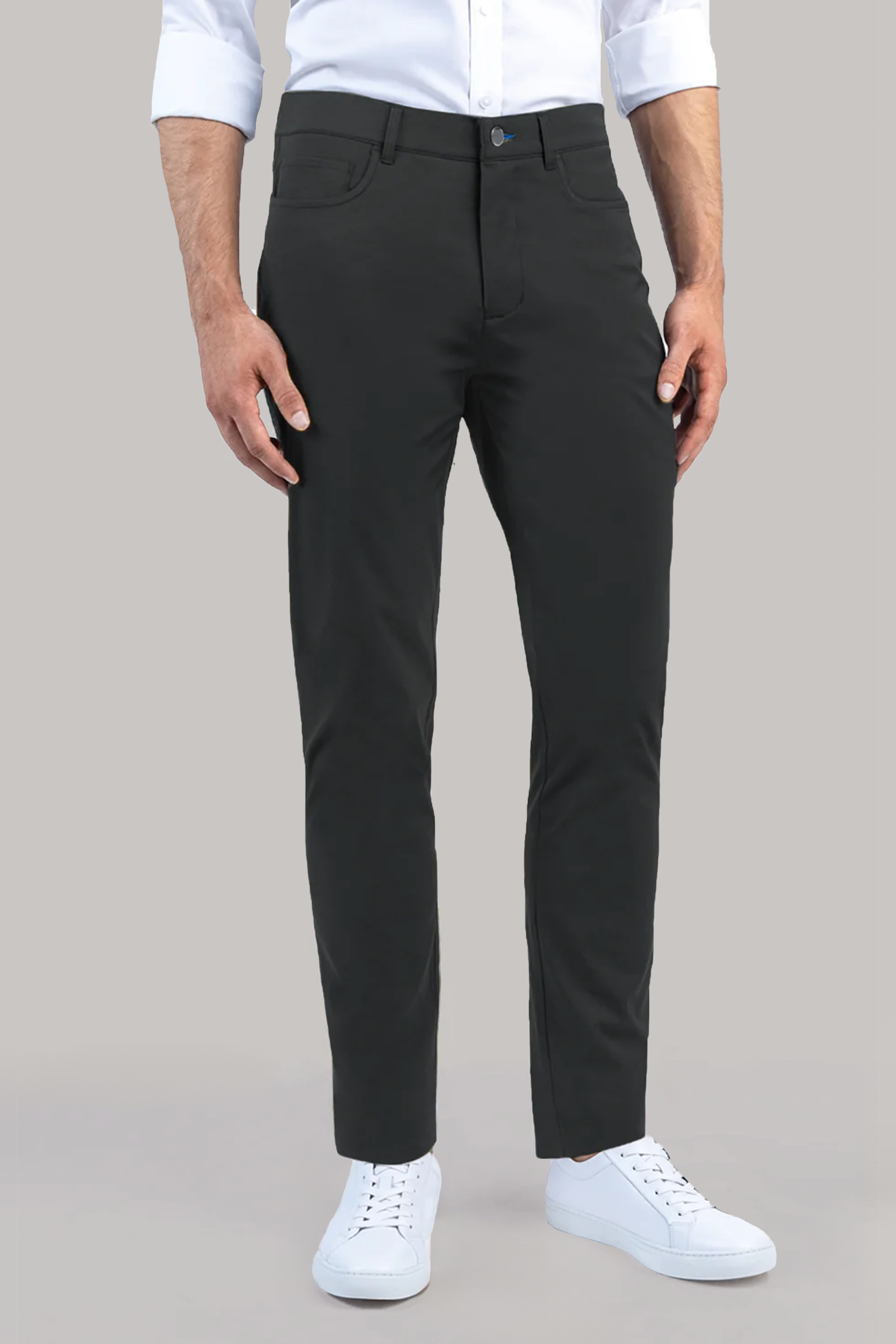 A man, head out of frame, stands against a neutral background wearing Twillory's Performance Five Pocket pants in Charcoal with a white 4-way stretch shirt (sleeves rolled) and white sneakers. His hands are relaxed at his sides.