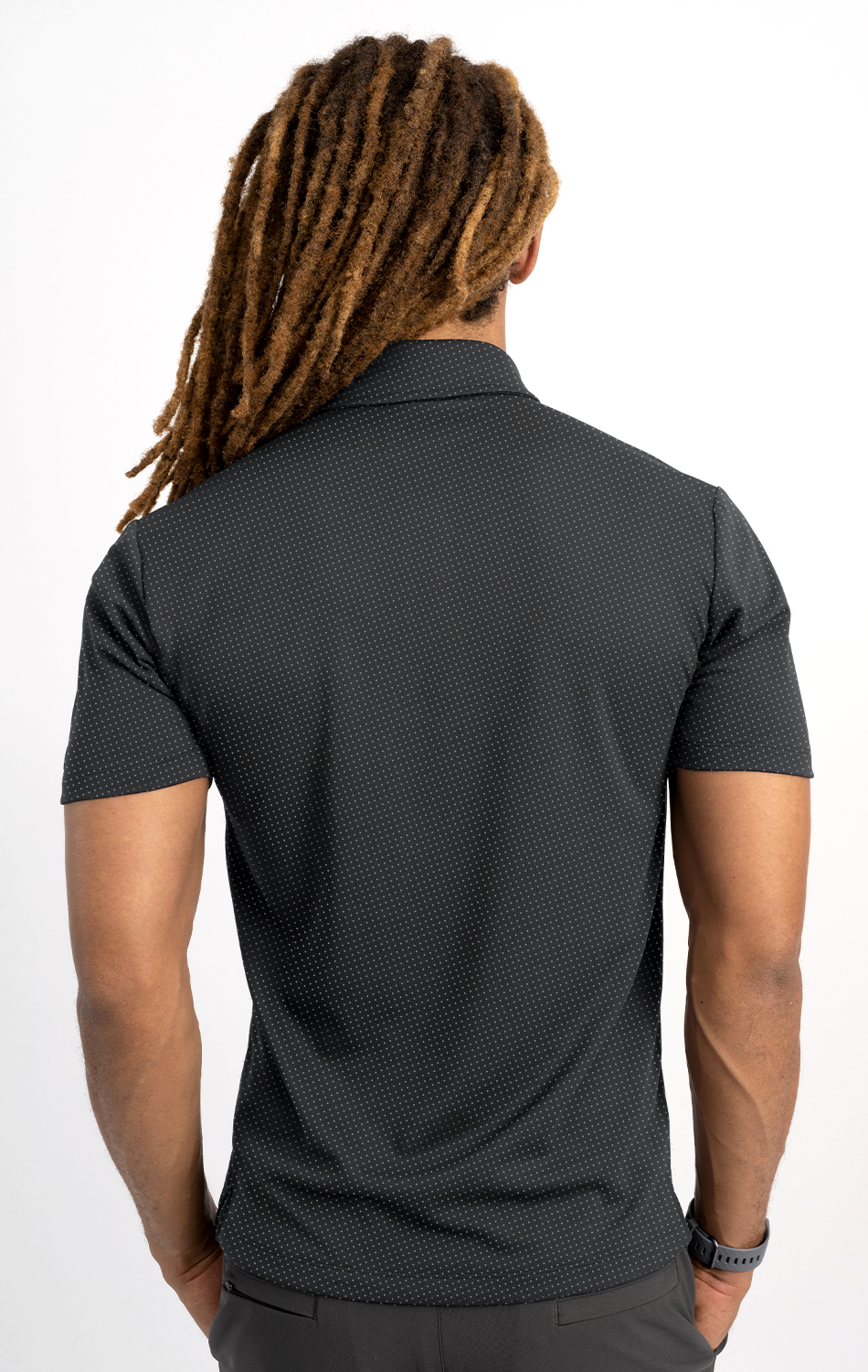 A person with long, thick brown dreadlocks faces away, wearing the Twillory Short Sleeve Polo Dot in Charcoal and dark pants, arms at their sides against a plain white background.