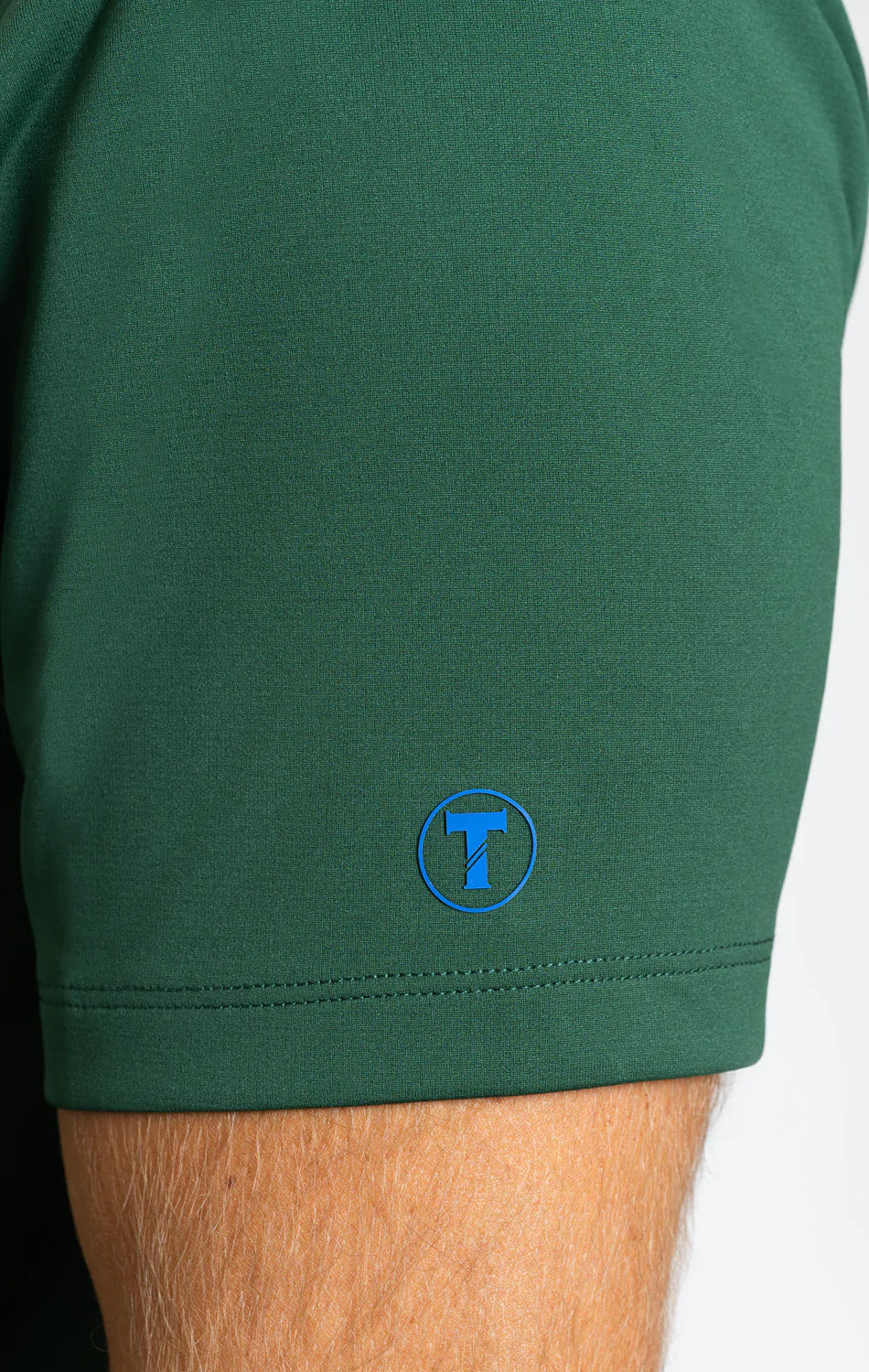 Close-up of a person’s arm wearing the Twillory Short Sleeve Polo Solid in Forest. The sleeve edge displays a small, circular blue Twillory "T" logo near the hem. The neutral background is softly blurred.