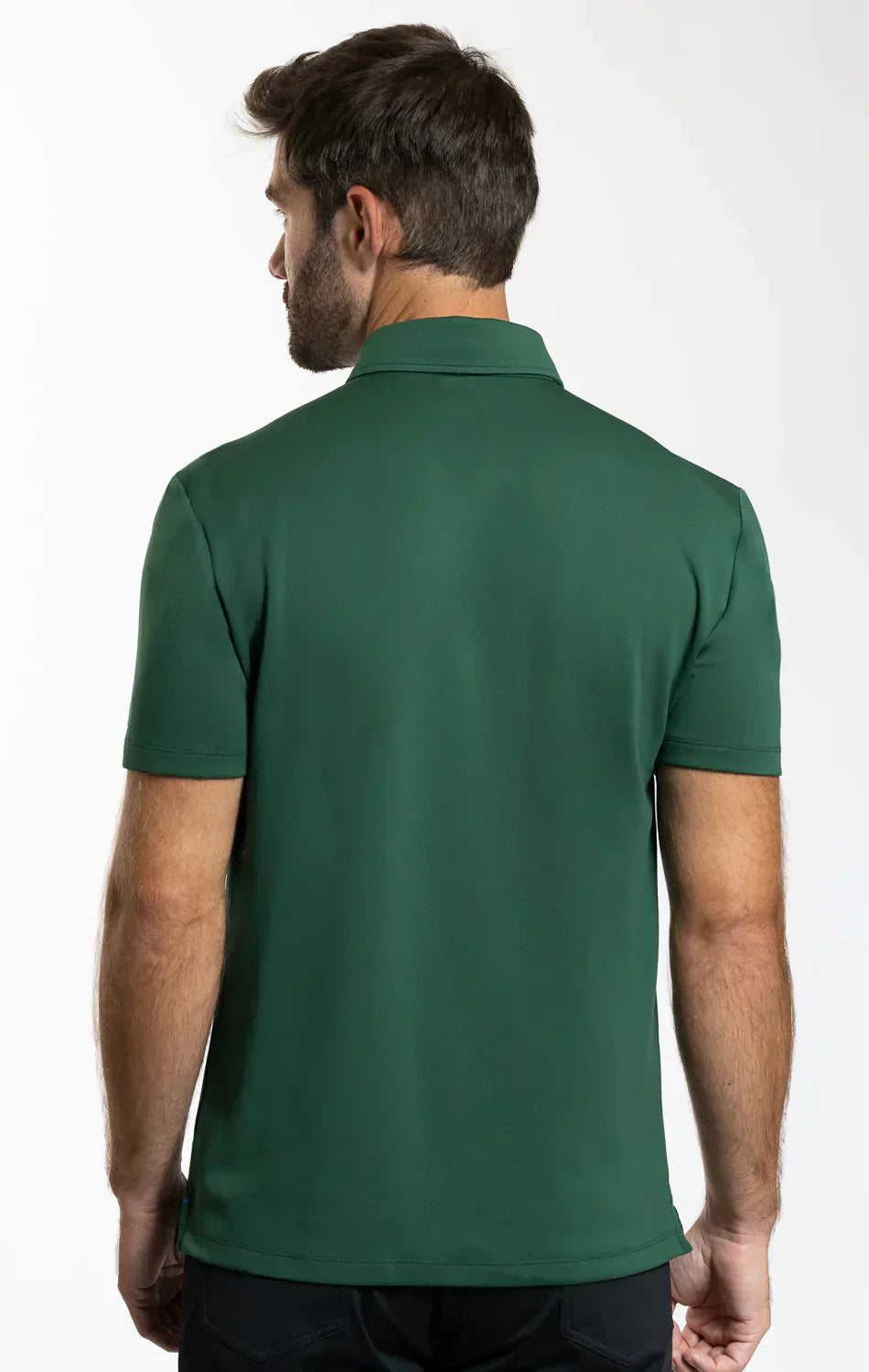 A man with short brown hair and a beard faces away from the camera, wearing the Twillory Short Sleeve Polo Solid in Forest and dark pants. He stands against a plain white background. The polo features moisture-wicking fabric.