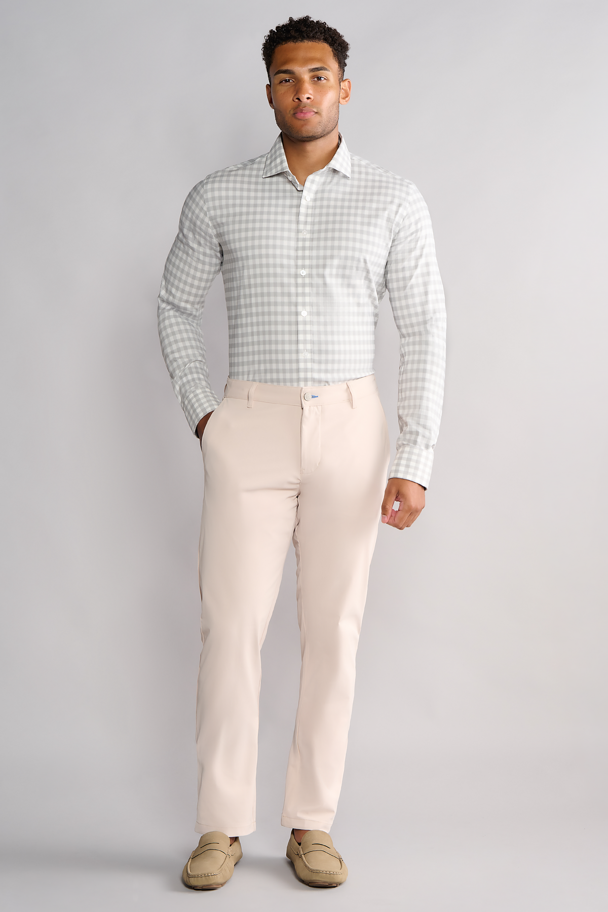 A man stands against a light gray background, wearing Twillory’s Leader Gingham // Grey White Performance Shirt tucked into slim, light beige dress pants. He completes the modern look with light tan loafers and one hand in his pocket.