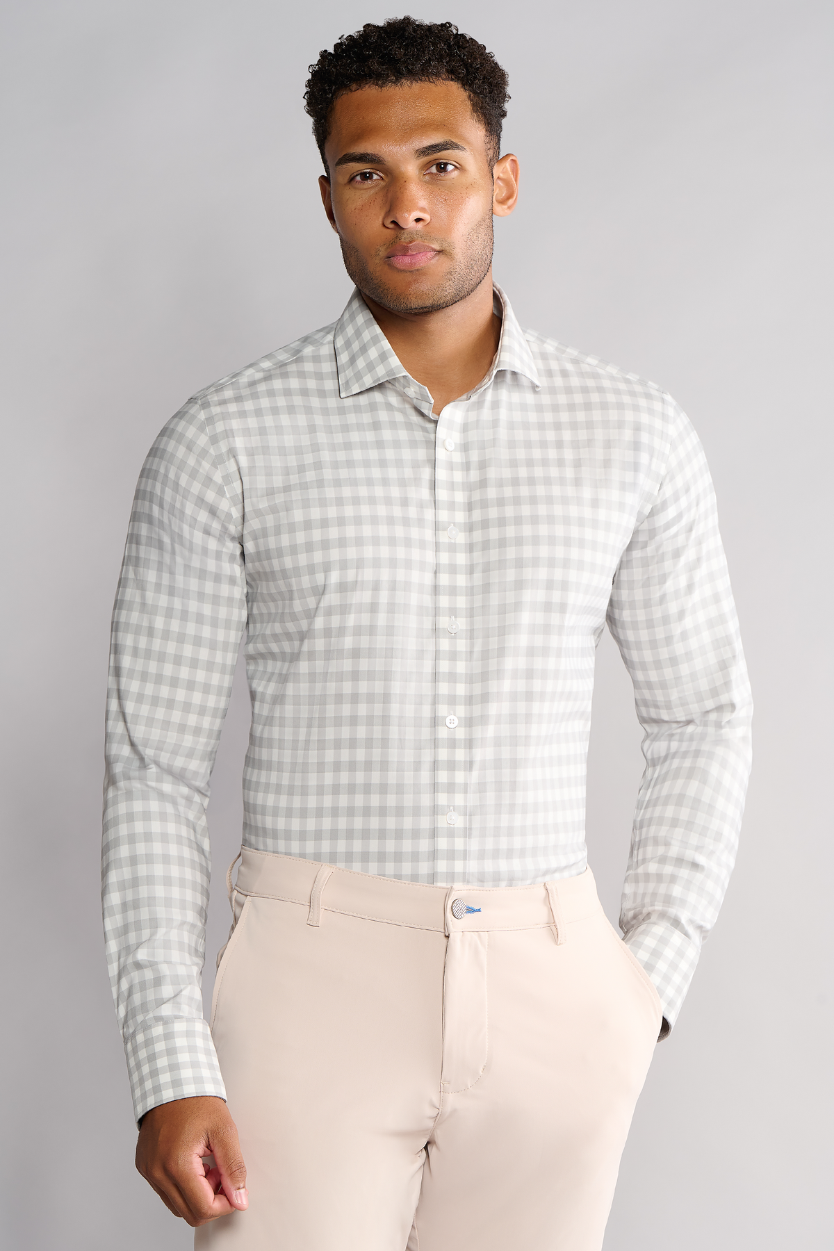 A man with curly hair wears the Twillory Leader Gingham // Grey White shirt tucked into beige pants, standing against a plain light gray background with one hand in his pocket and a neutral expression.