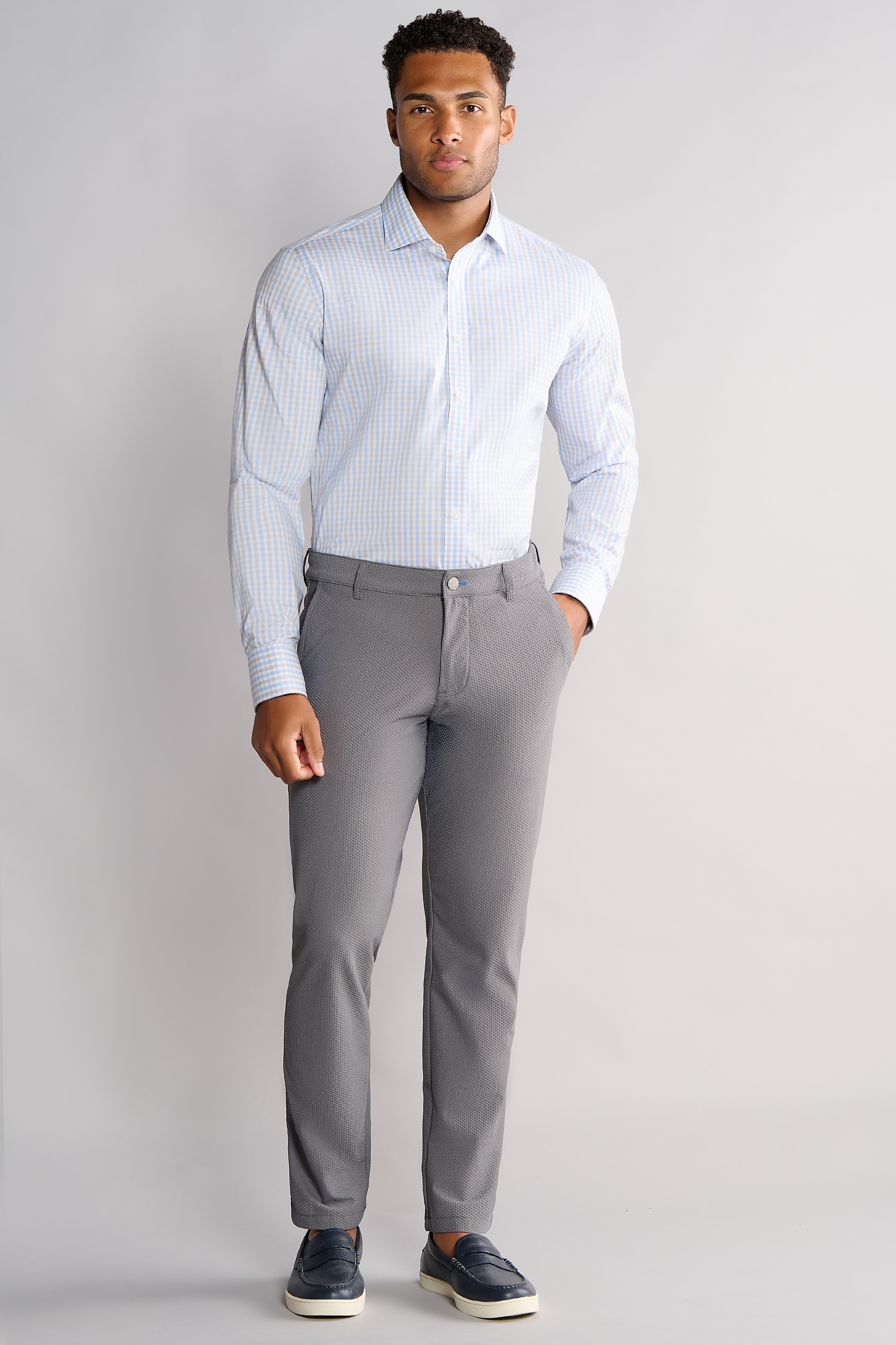 A man stands facing forward against a light gray background, wearing Twillory's Leader Check // Grey Blue Tattersall performance shirt, tucked into gray slim-fit trousers, paired with dark blue loafers with white soles.