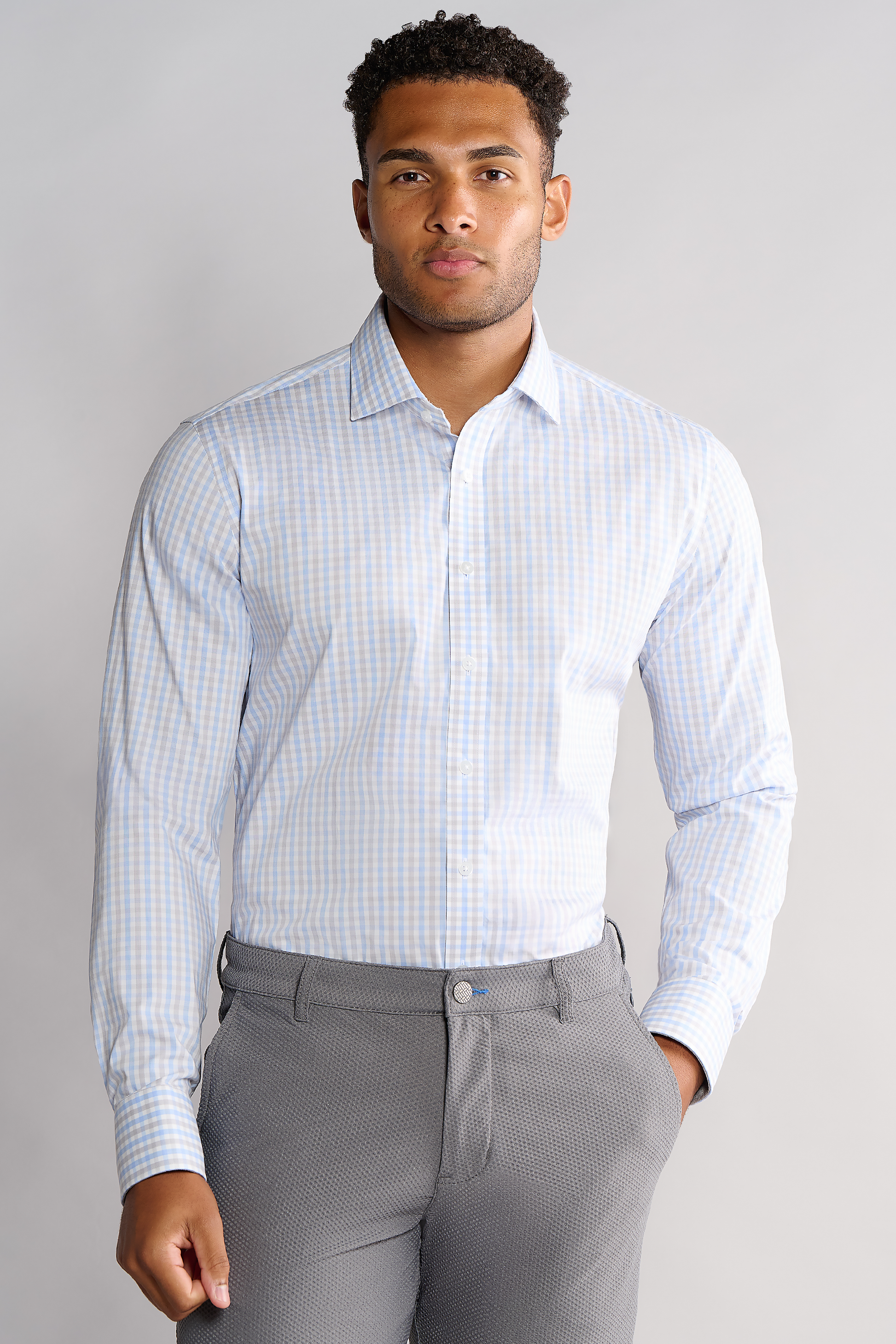 A man with short curly hair and a neutral expression faces forward, wearing Twillory's Leader Check // Grey Blue Tattersall shirt tucked into light gray dress pants. His right hand rests at his side against a plain, light-colored background.