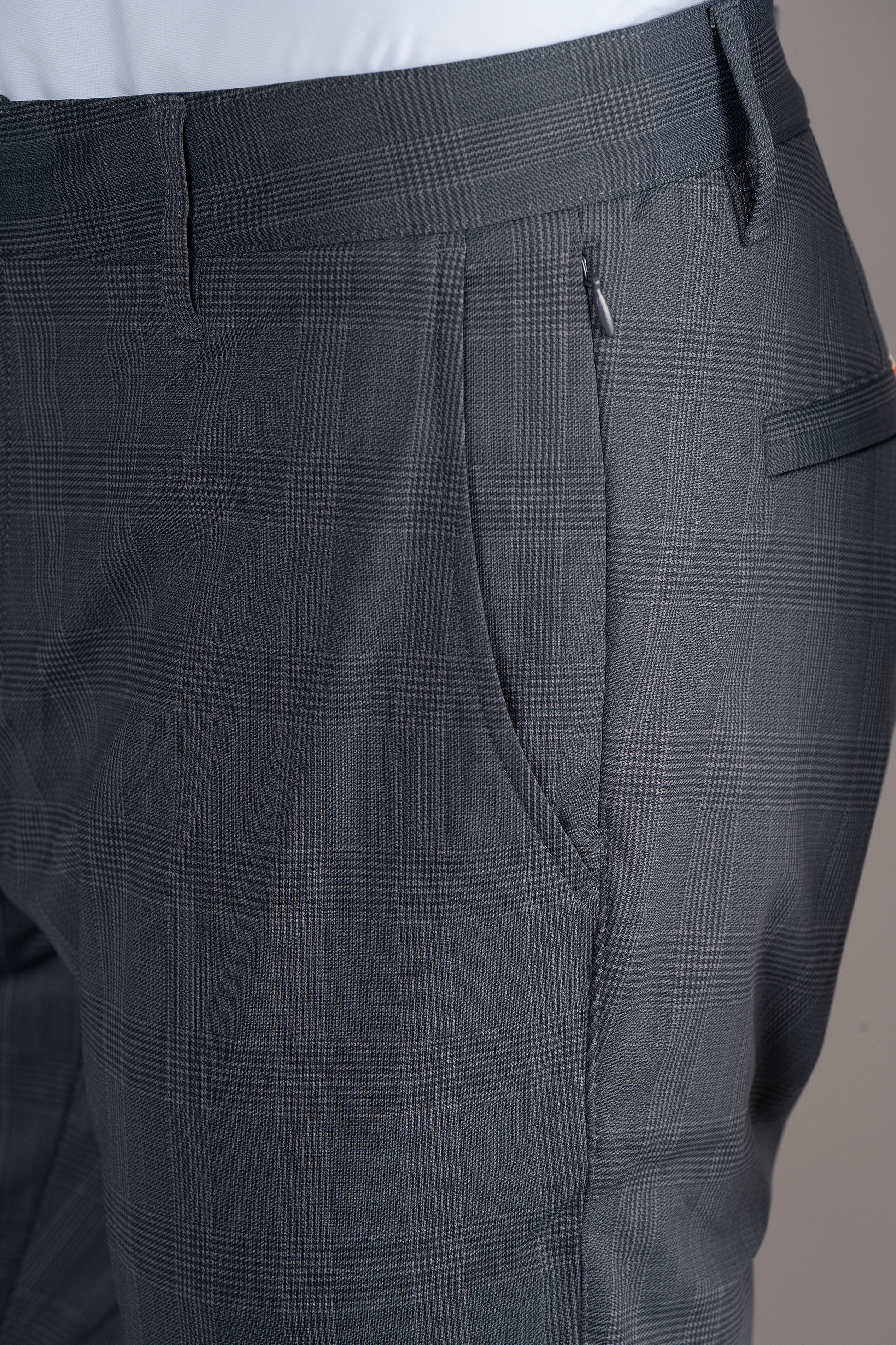 Close-up of a person wearing Twillory Performance Pants // Grey Prince of Wales, showcasing the subtle checked pattern, waistband with belt loops, pocket, and zipper pull against a plain neutral background.