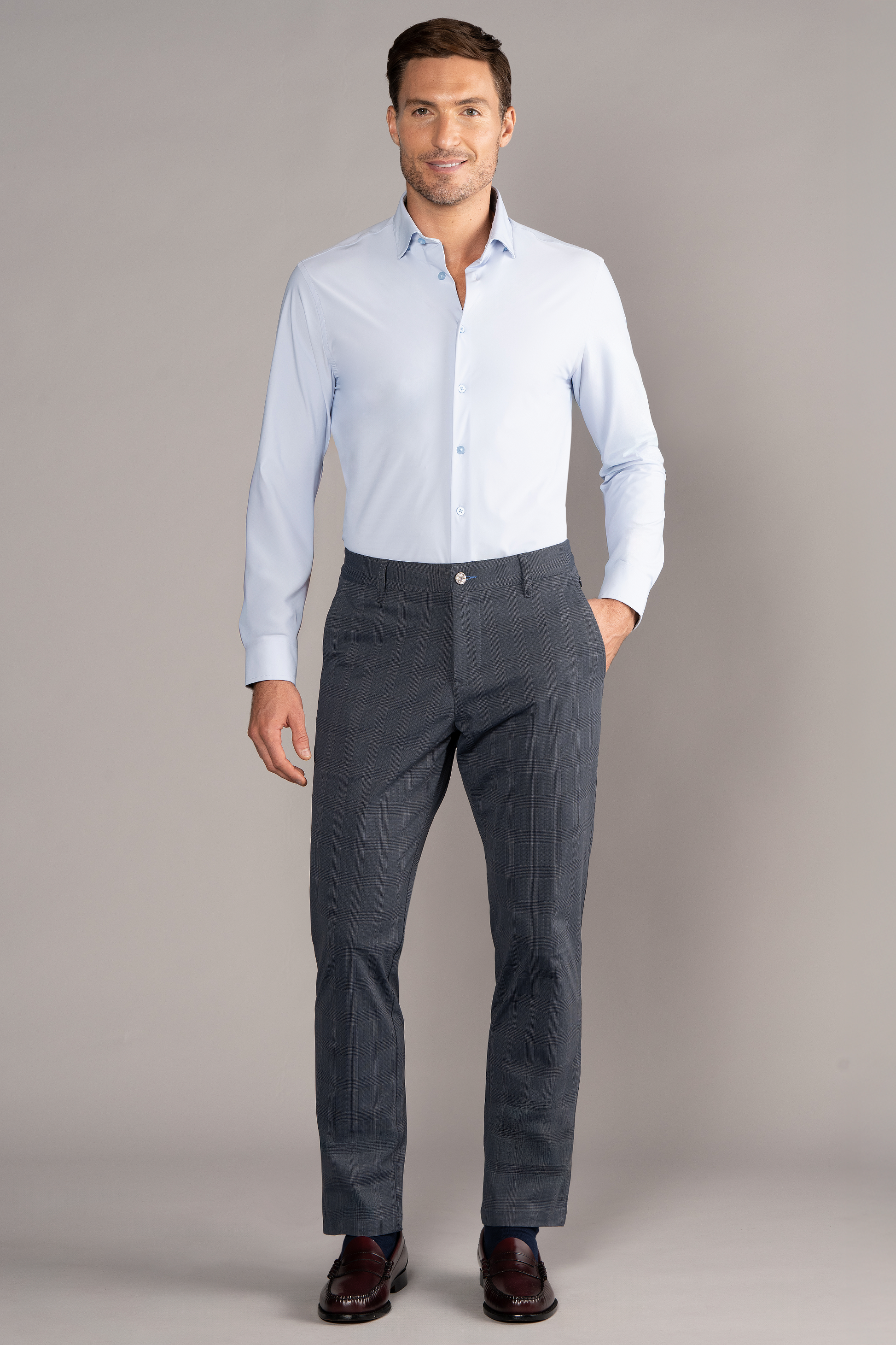 A man stands against a plain gray background, smiling. He wears a light blue button-up shirt tucked into Twillory Performance Pants in Grey Prince of Wales, with his left hand in his pocket and wearing dark brown dress shoes.