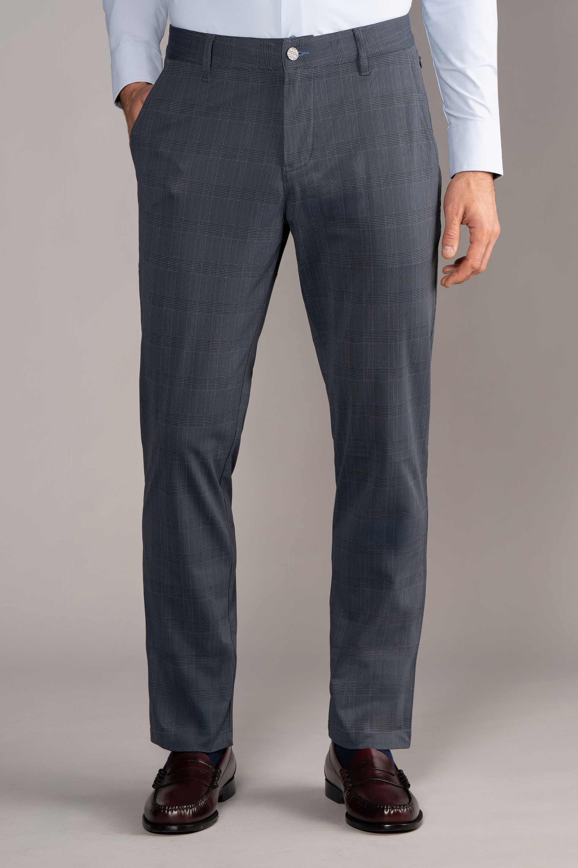 A man wears Twillory’s Performance Pants in Grey Prince of Wales, paired with a tucked-in light blue dress shirt and dark brown loafers. His hands are in his pockets, chest down visible, standing against a neutral background.