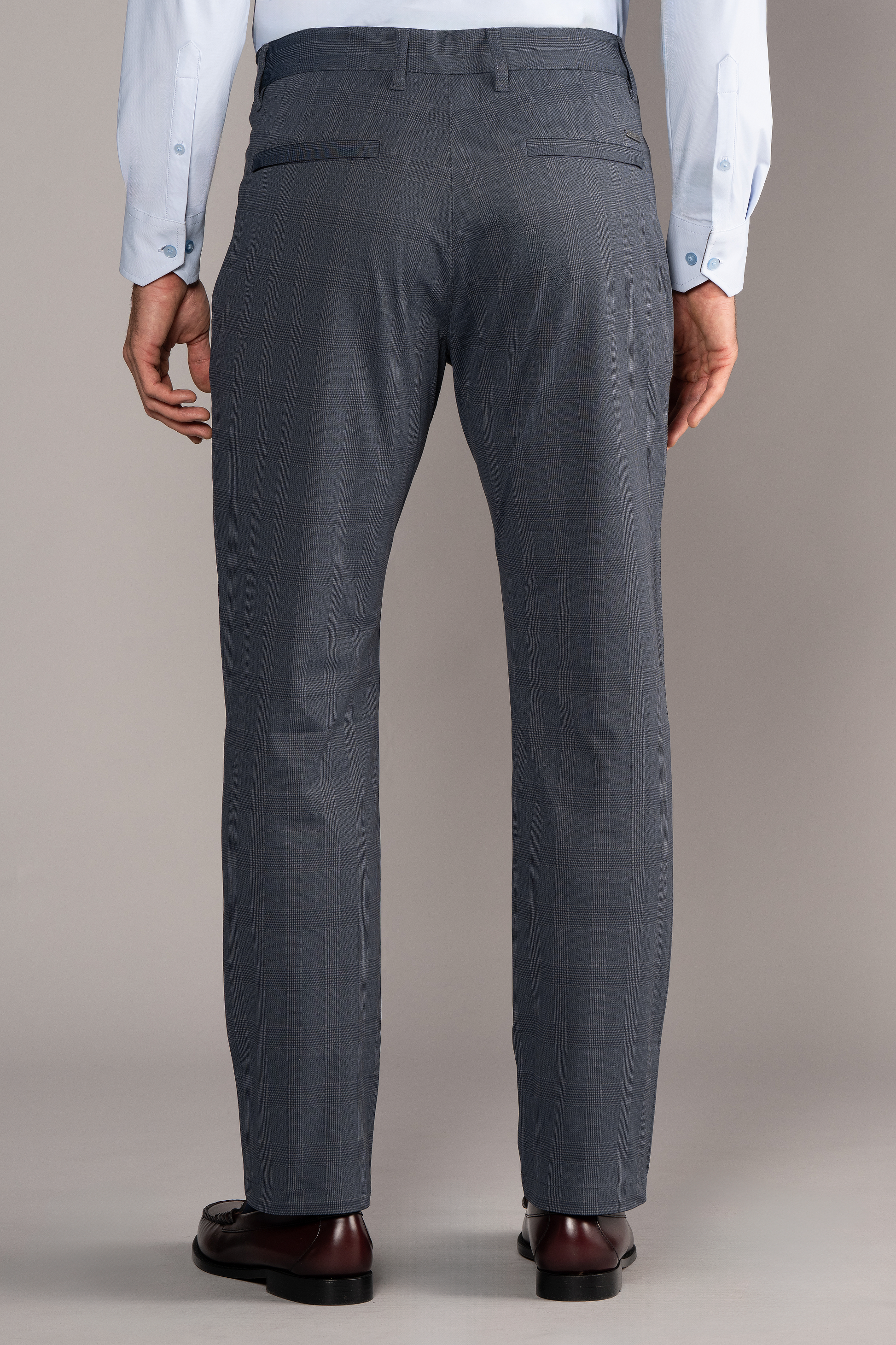 A man stands facing away, wearing Twillory Performance Pants // Grey Prince of Wales and a light blue shirt. His arms rest by his sides, with a neutral background highlighting the stretch fabric and modern fit. Image is cropped above his shoulders.
