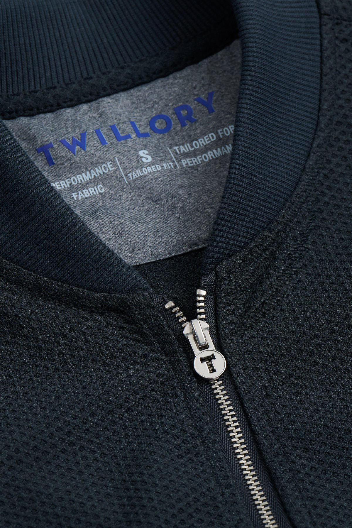 Close-up of the Twillory AIR Bomber // Heather Charcoal jacket, featuring a dark textured fabric, partially zipped silver zipper, and a blue-on-gray label noting performance fabric and breathable comfort in size S.