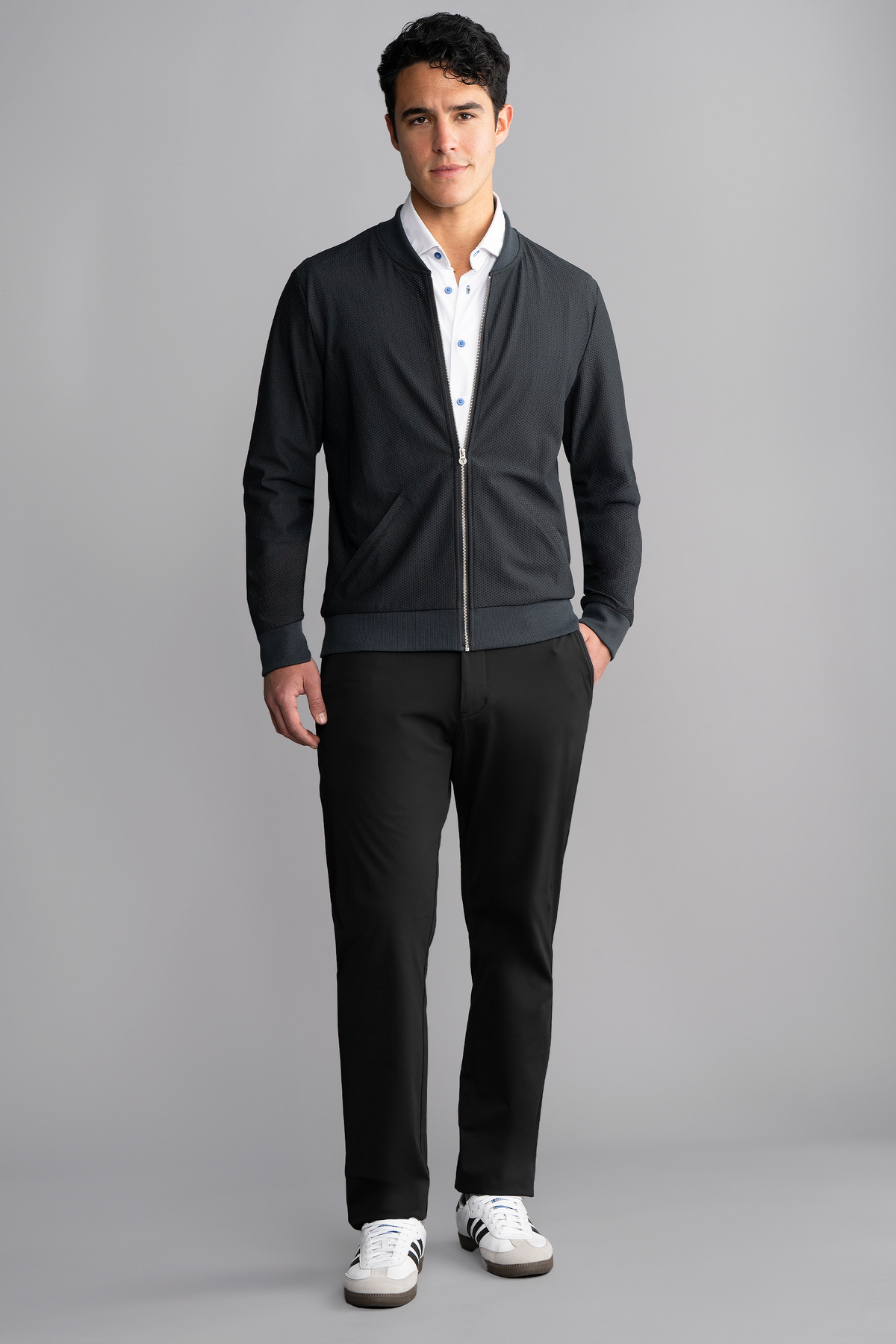 A man with short dark hair stands against a gray background, wearing a Twillory AIR Bomber // Heather Charcoal—crafted with breathable 360° Airflow Fabric—over a white shirt, black pants, and white sneakers. He looks at the camera neutrally.