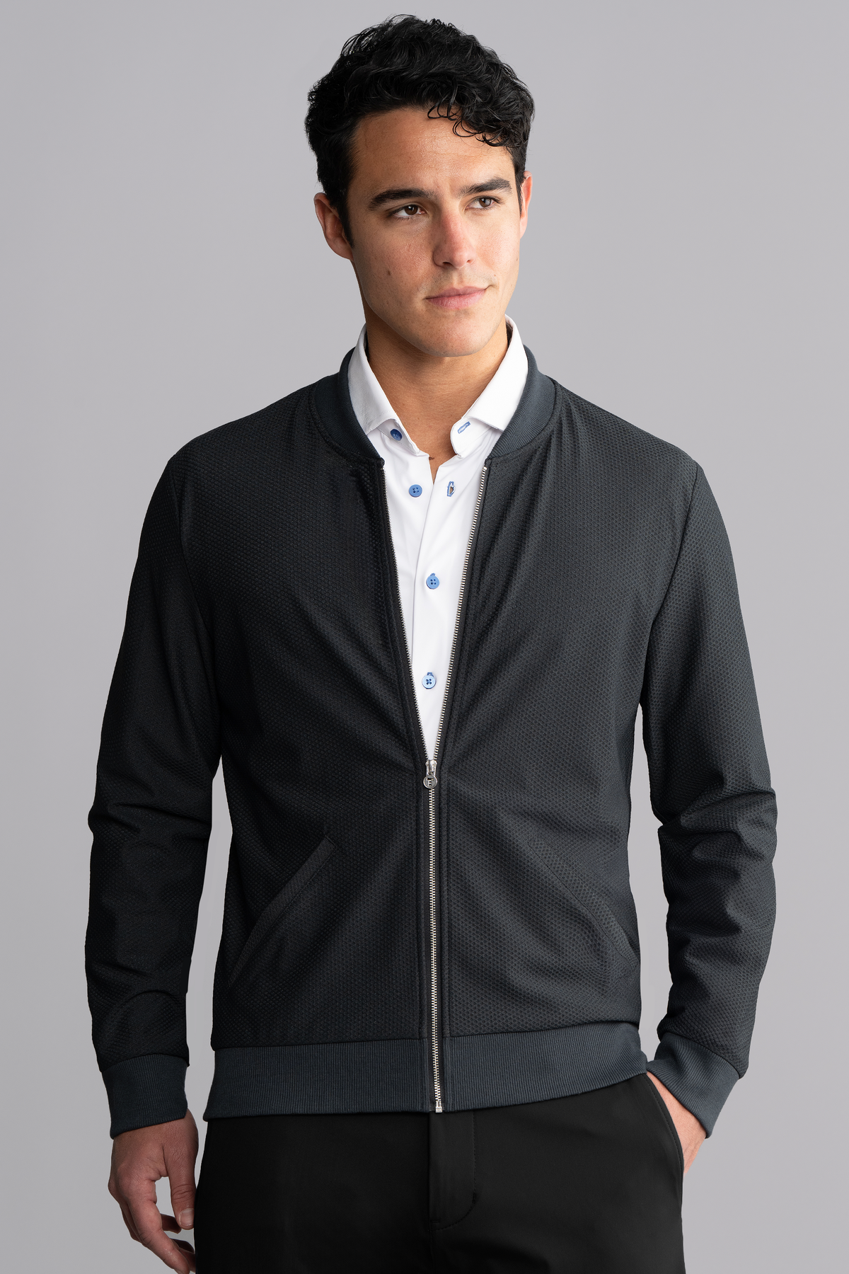 A young man with short curly dark hair wears the Twillory AIR Bomber // Heather Charcoal over a white dress shirt with blue buttons, standing against a plain light grey background and looking slightly left with a neutral expression.