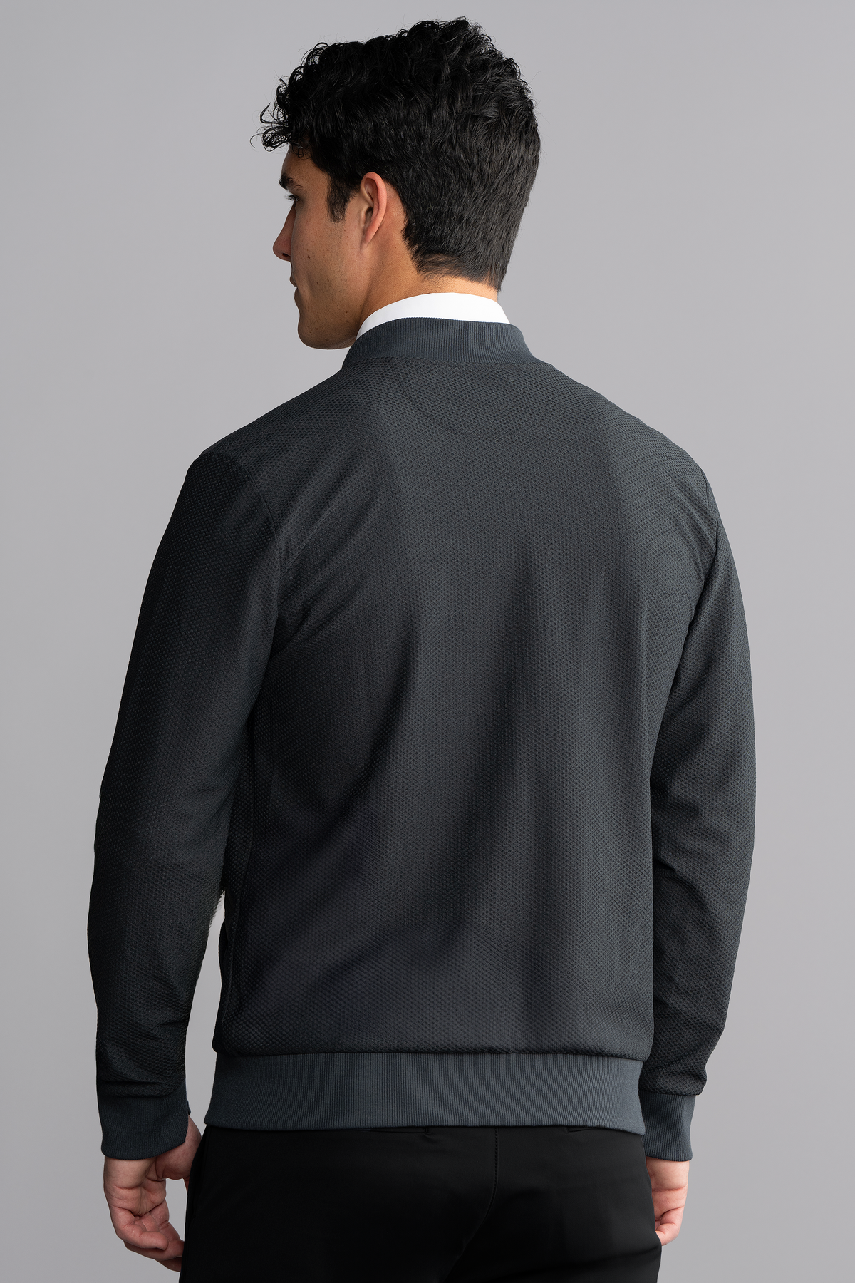 A man with short, dark hair stands with his back to the camera against a gray background, wearing Twillory's AIR Bomber // Heather Charcoal made from 360° Airflow Fabric with ribbed details, paired with black pants. His head is turned slightly left.