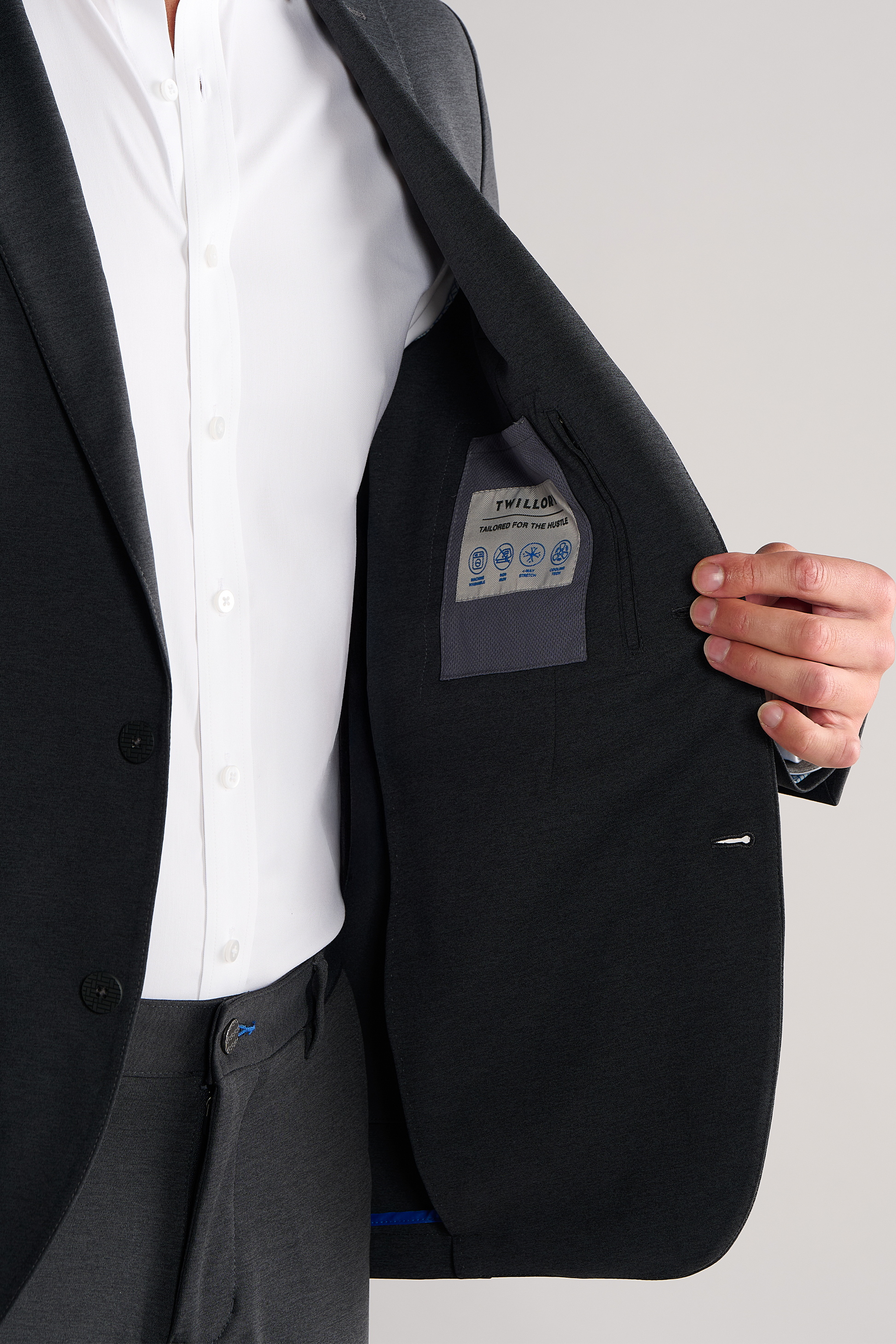 A person in a dark suit and white shirt holds open their Circular Knit Blazer // Heather Grey by Twillory to show the inner lining and care label. Only the torso and hand are visible, with a plain background highlighting this blazer’s thoughtful details.
