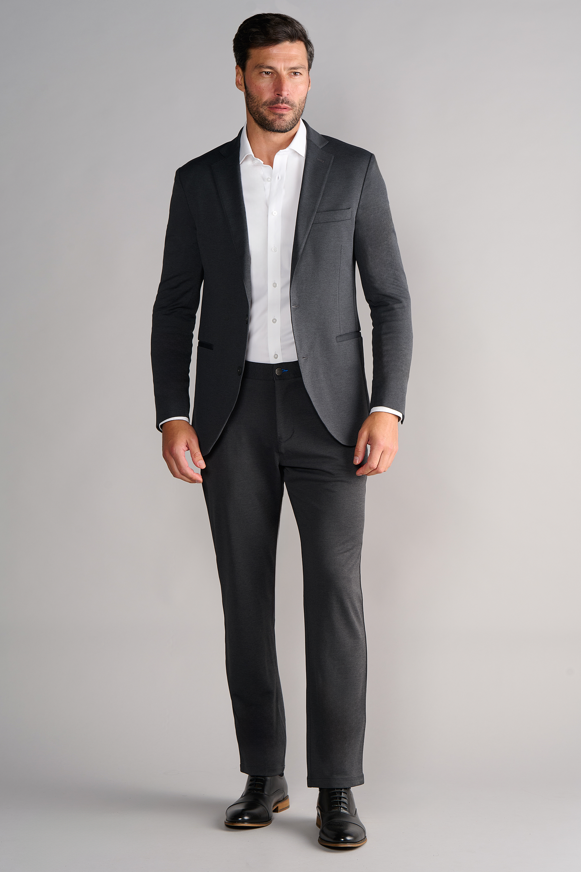 A man with short dark hair and a trimmed beard stands against a plain light gray background, wearing Twillory's Circular Knit Blazer in Heather Grey over a white dress shirt and black dress shoes. His jacket is buttoned, hands relaxed at his sides.