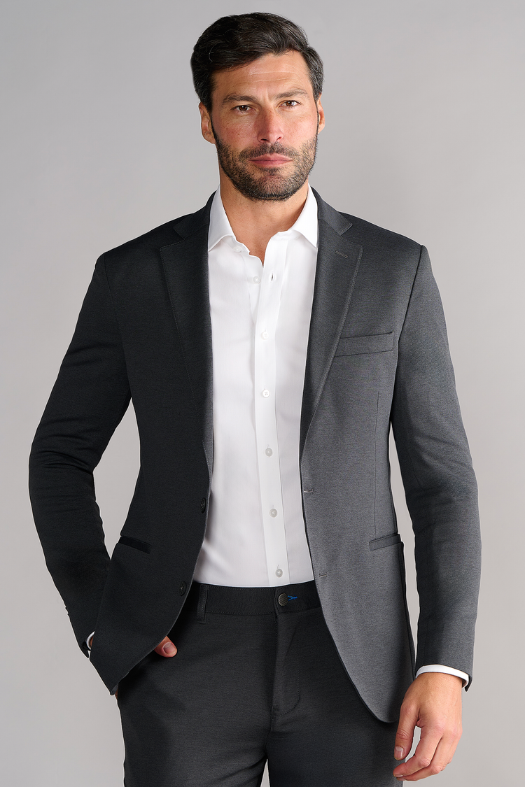 A man with dark hair and a beard wears the Twillory Circular Knit Blazer in Heather Grey over a white dress shirt. He stands facing forward with one hand in his pocket against a light gray background, showing a neutral expression.