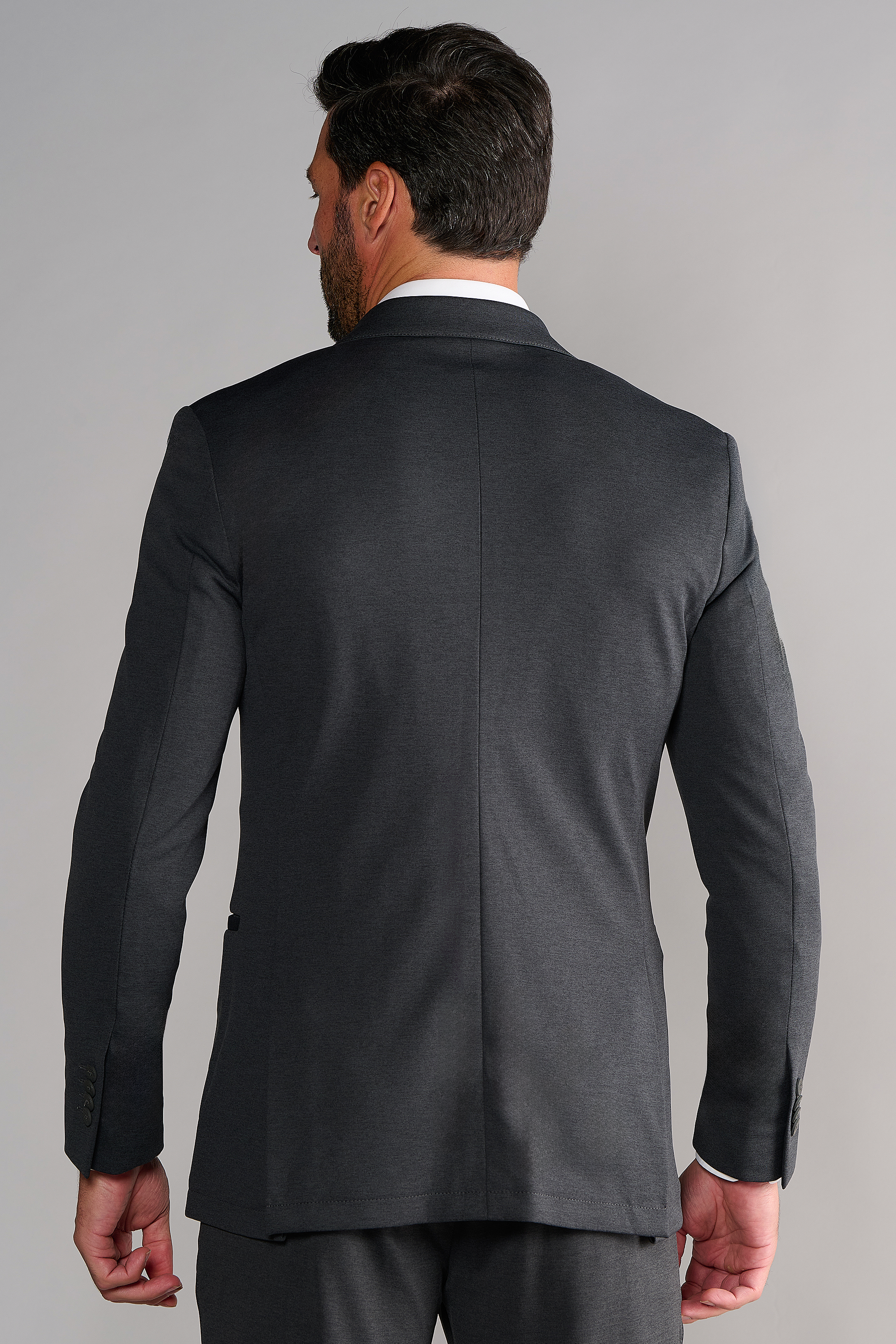 A man with dark hair and a short beard is seen from behind, wearing the Twillory Circular Knit Blazer in Heather Grey. His right hand rests at his side against a plain, light gray background.