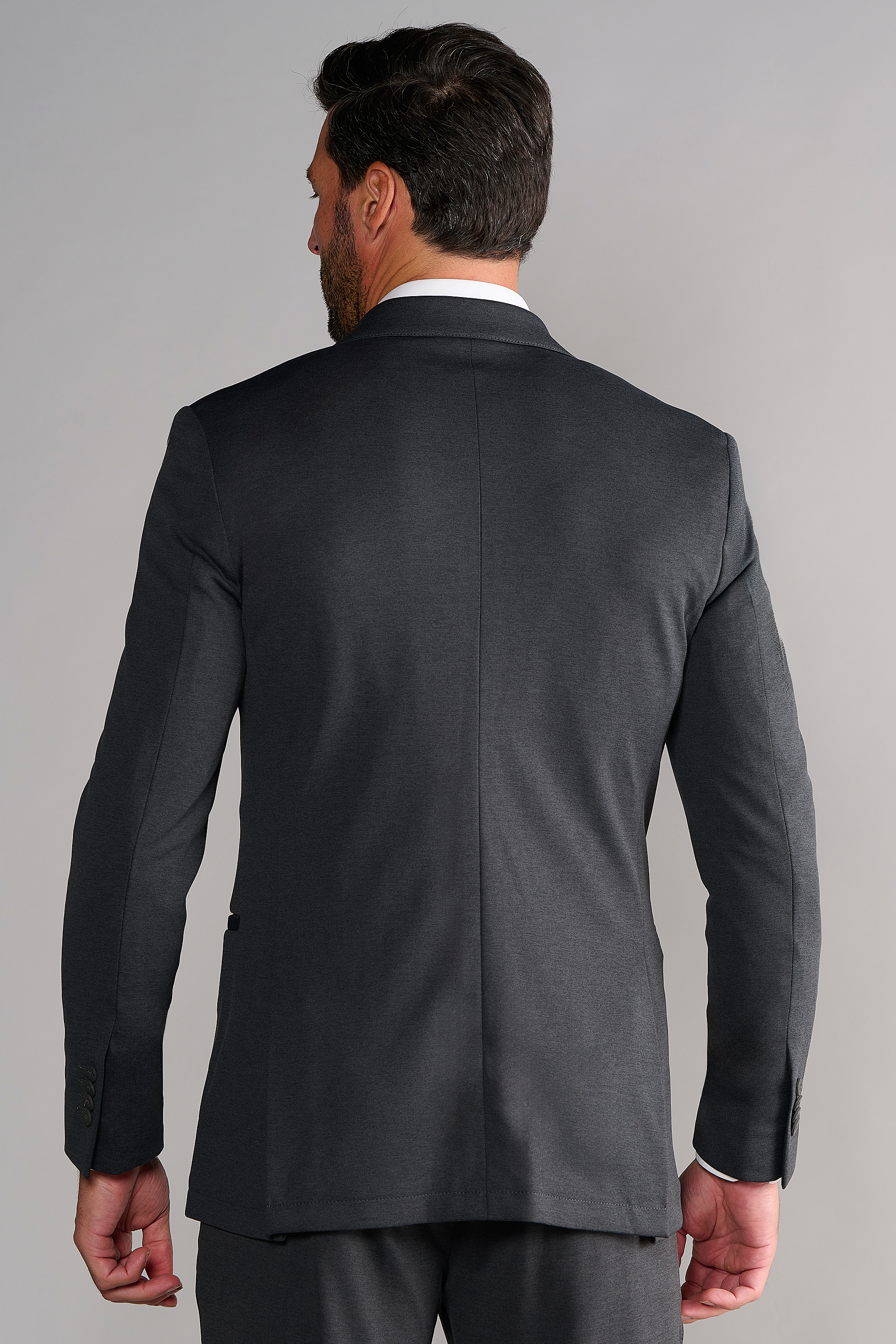 A man with dark hair and a short beard is seen from behind, wearing the Twillory Circular Knit Blazer in Heather Grey. His right hand rests at his side against a plain, light gray background.