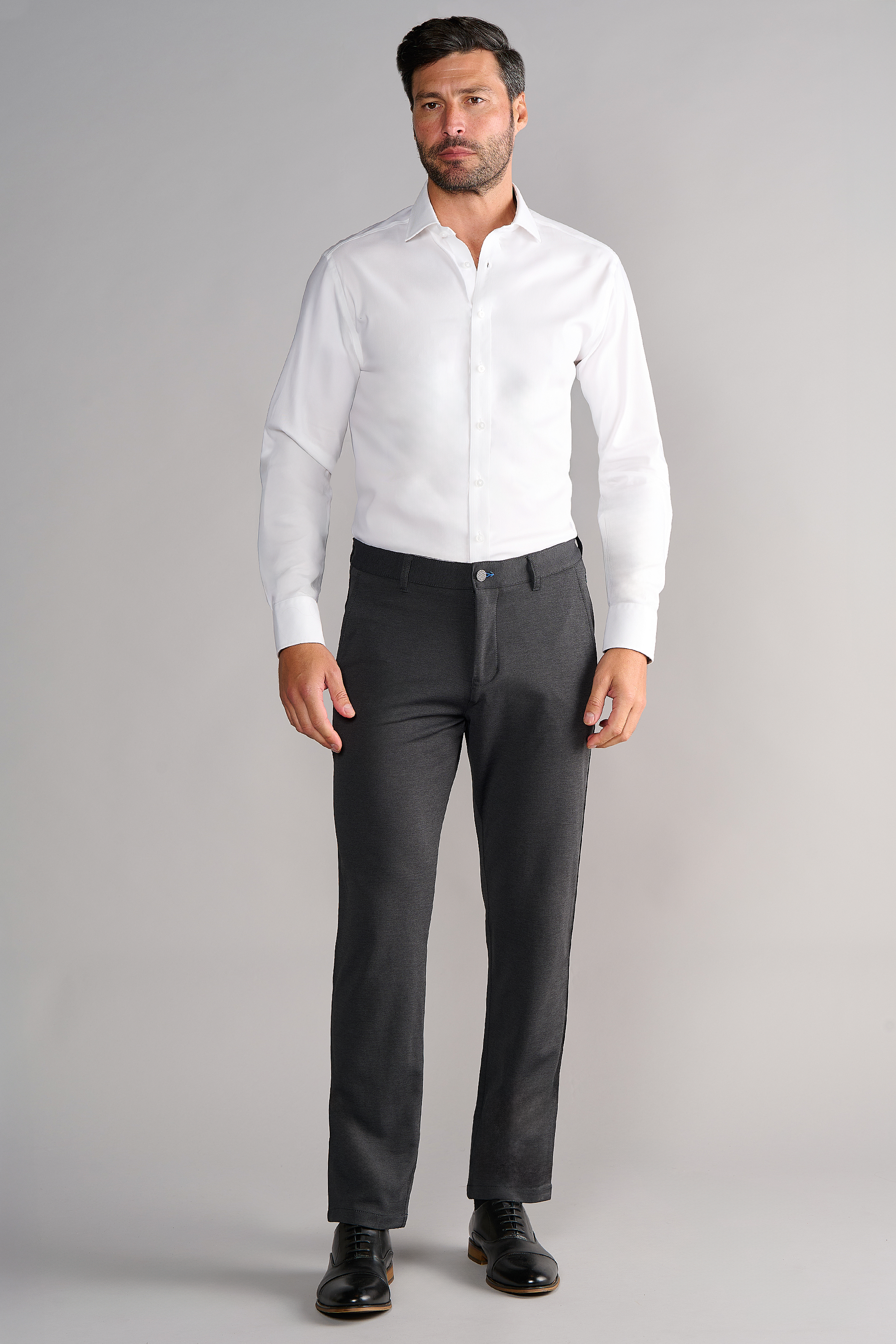 A man with short dark hair, light skin, and a beard stands against a light gray background wearing Twillory Circular Knit Pants in Heather Grey, a fitted white dress shirt, and black dress shoes.
