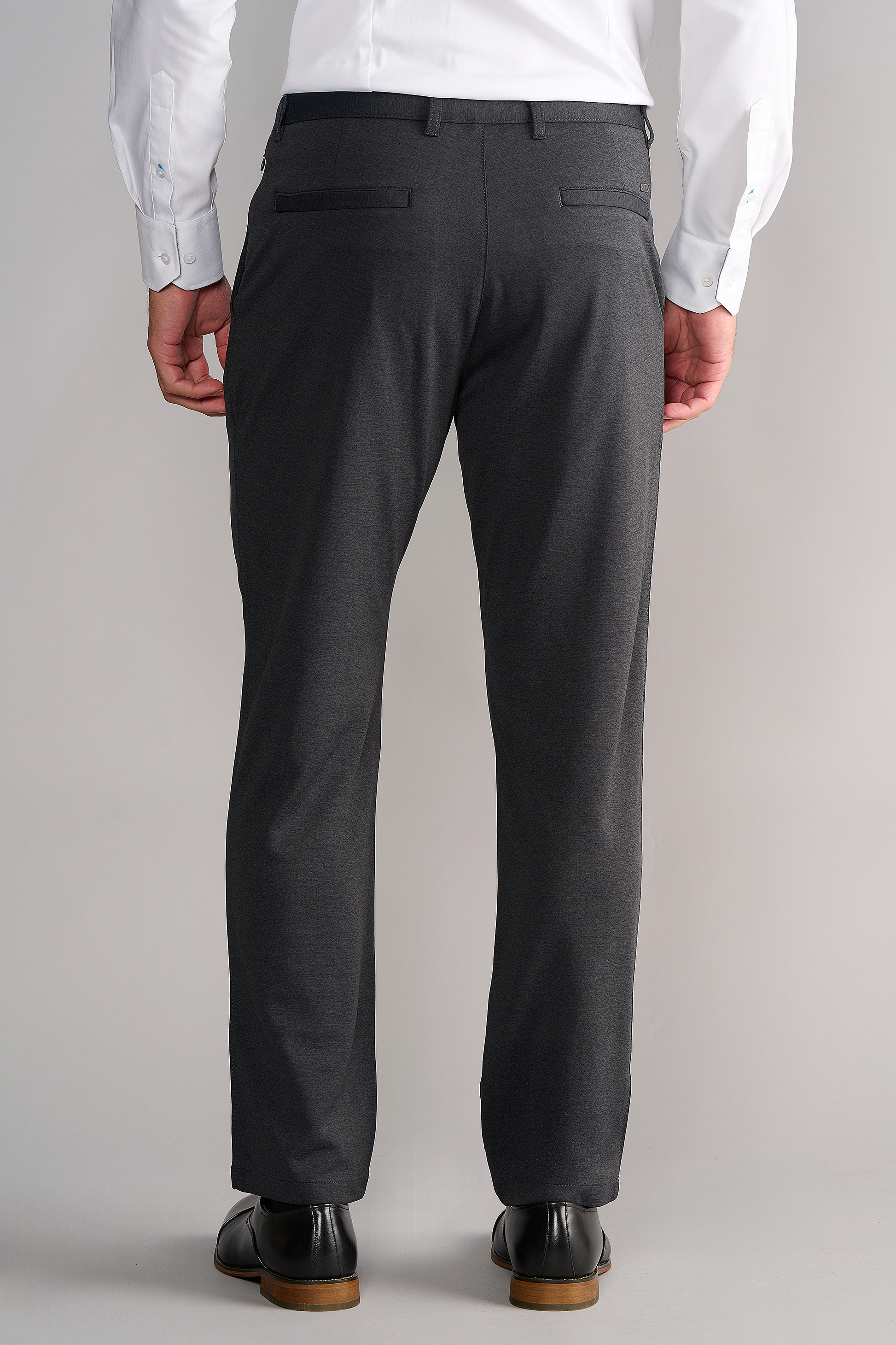 A person faces away from the camera, wearing Twillory Circular Knit Pants in Heather Grey with two back flap pockets and belt loops, a white long-sleeve shirt, and black dress shoes against a plain light background.