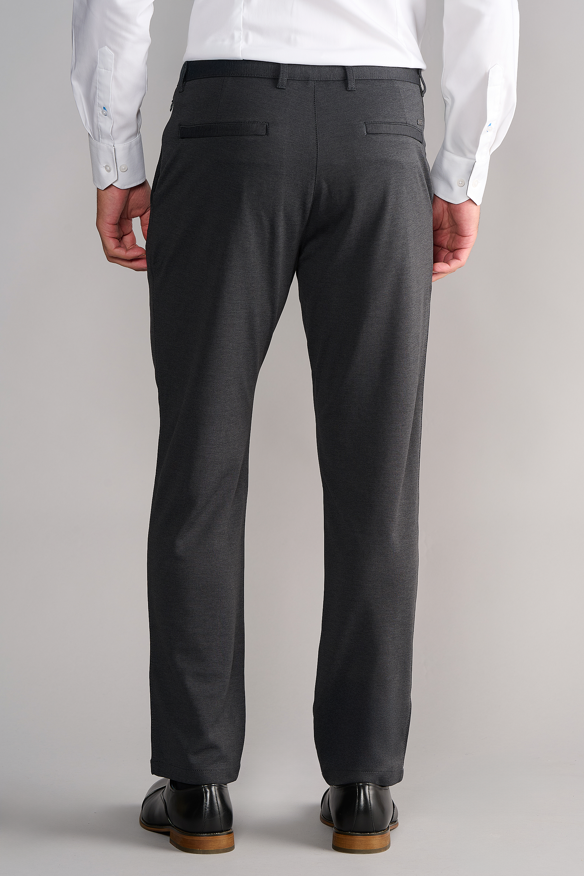 A person faces away from the camera, wearing Twillory Circular Knit Pants in Heather Grey with two back flap pockets and belt loops, a white long-sleeve shirt, and black dress shoes against a plain light background.
