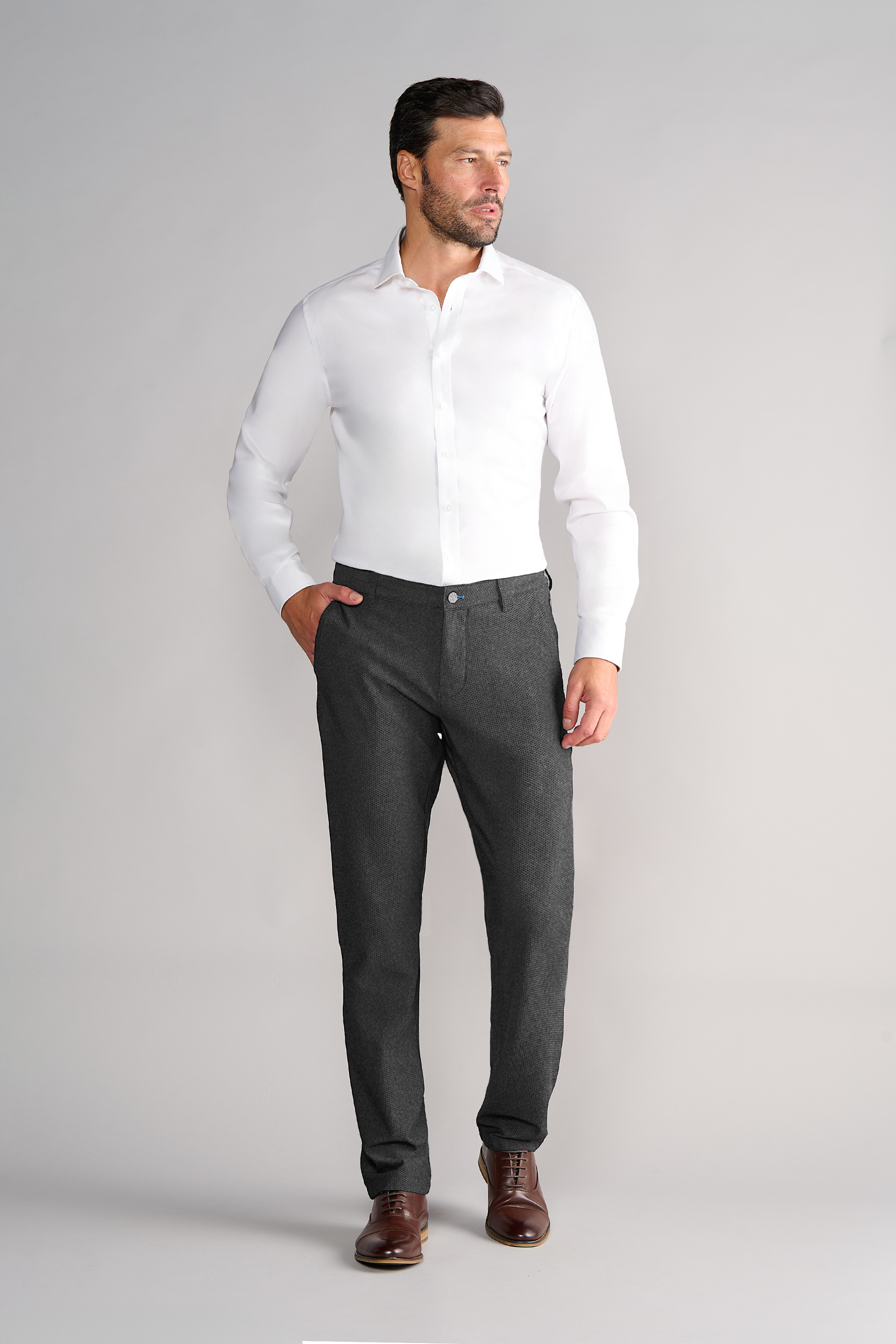 A man with short dark hair and a beard stands against a light gray background, wearing a white dress shirt, brown shoes, and Twillory AIR Pants in Heather Grey. One hand is in his pocket as he looks off to the side.