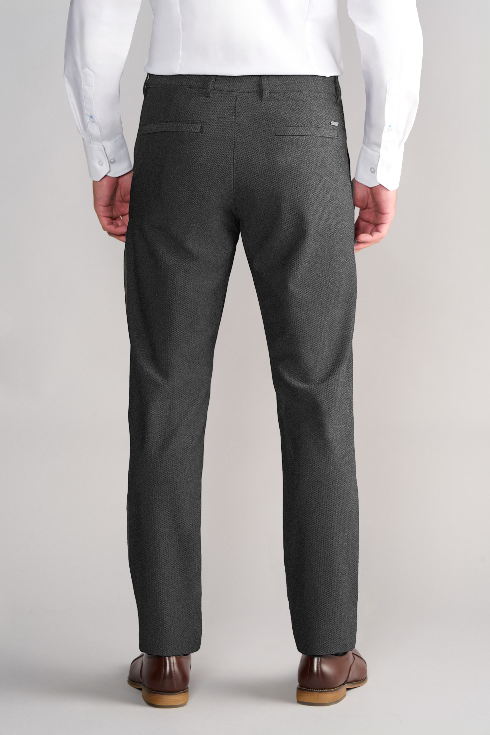 A man stands facing away, wearing a white dress shirt, brown dress shoes, and Twillory AIR Pants in Heather Grey with two back welt pockets. His arms rest at his sides against a plain light background.