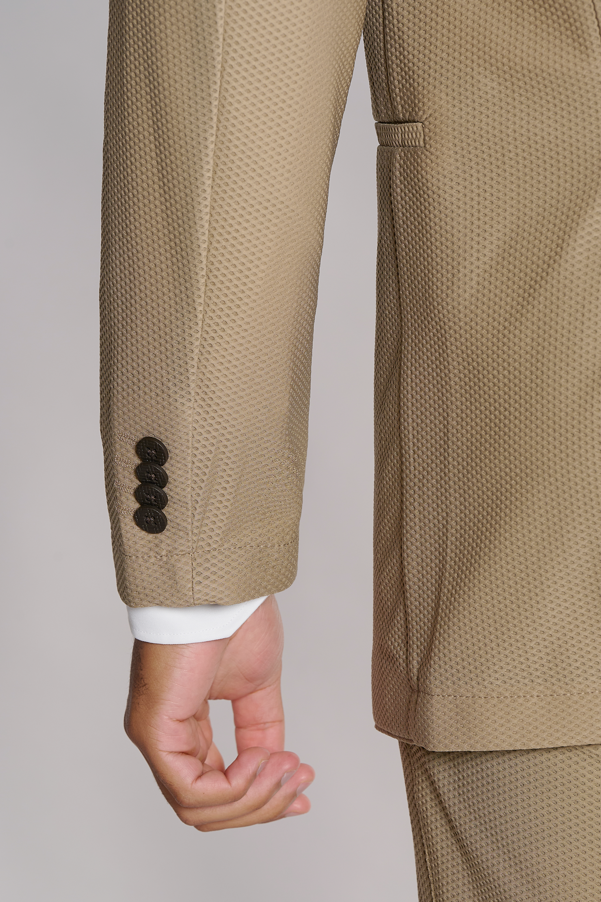 A close-up of a left arm in the Twillory AIR Blazer // Khaki shows its textured fabric and black sleeve buttons. The 360° airflow keeps you cool, while a relaxed hand and white cuff add style against a plain light background.