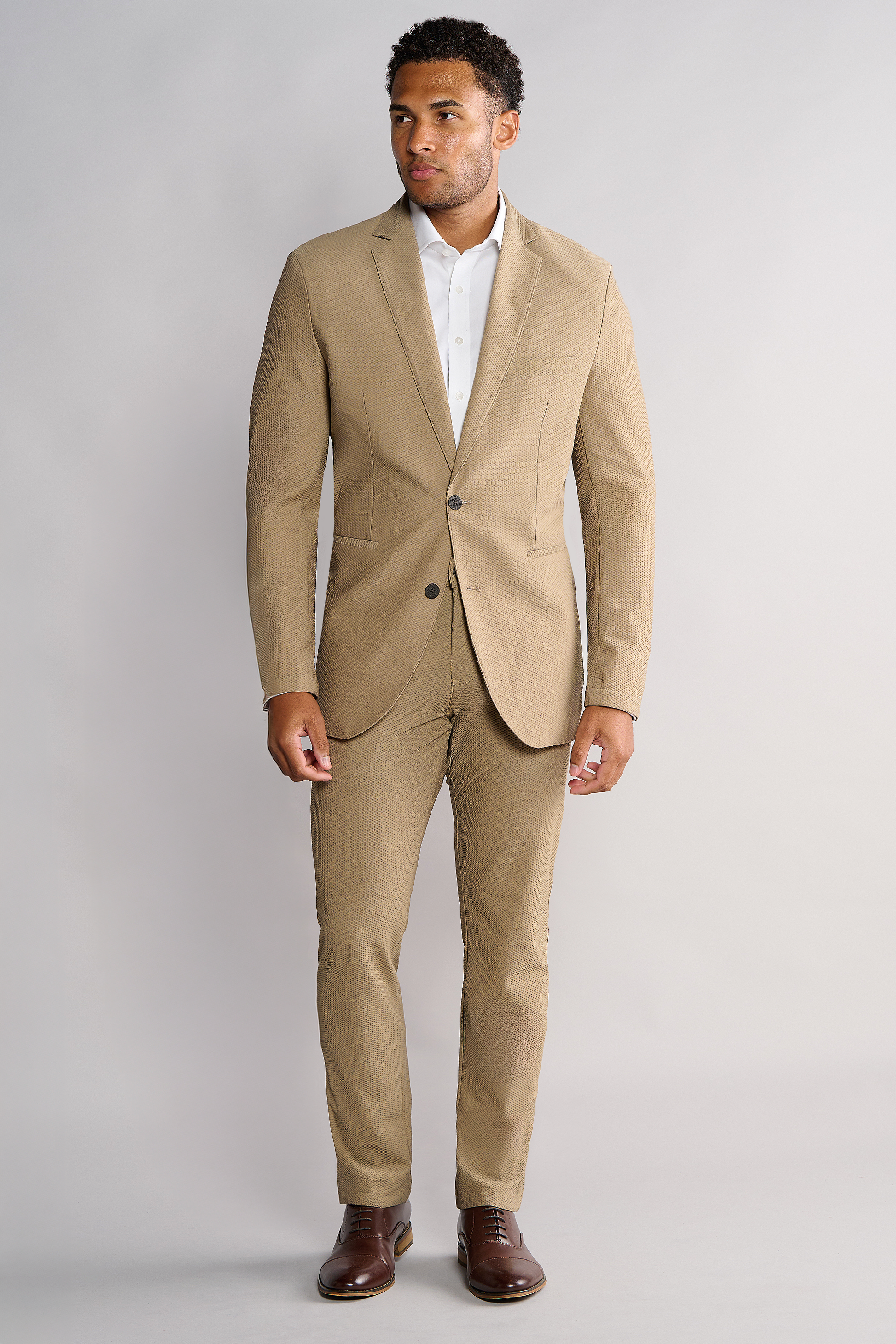 A man stands against a light gray background wearing the Twillory AIR Blazer // Khaki with 360° airflow fabric, paired with a white dress shirt and dark brown dress shoes. He looks slightly left, hands relaxed by his sides, with a neutral expression.