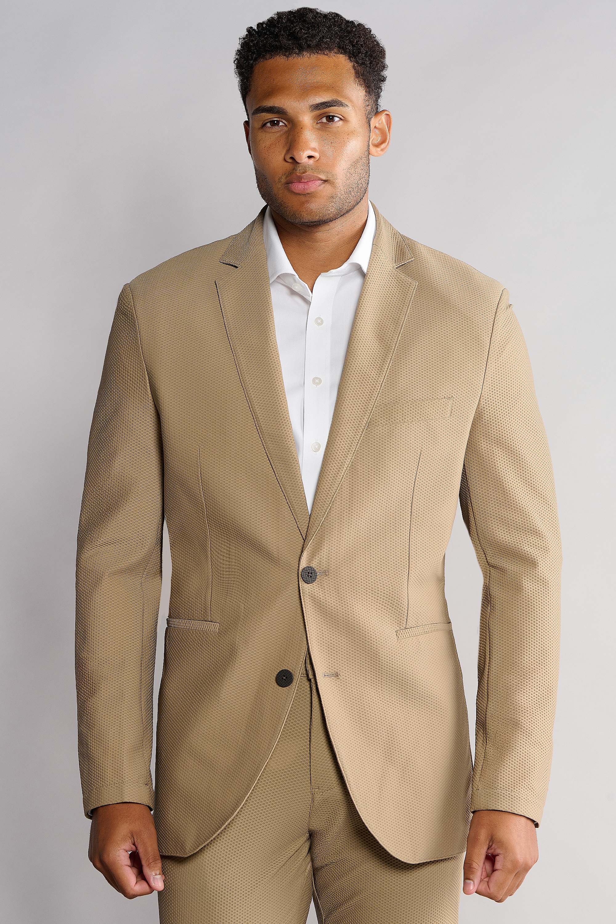 A man with short curly hair and a neutral expression stands against a light gray background, wearing Twillory’s AIR Blazer in Khaki—textured, machine washable, single-buttoned—over a white dress shirt.
